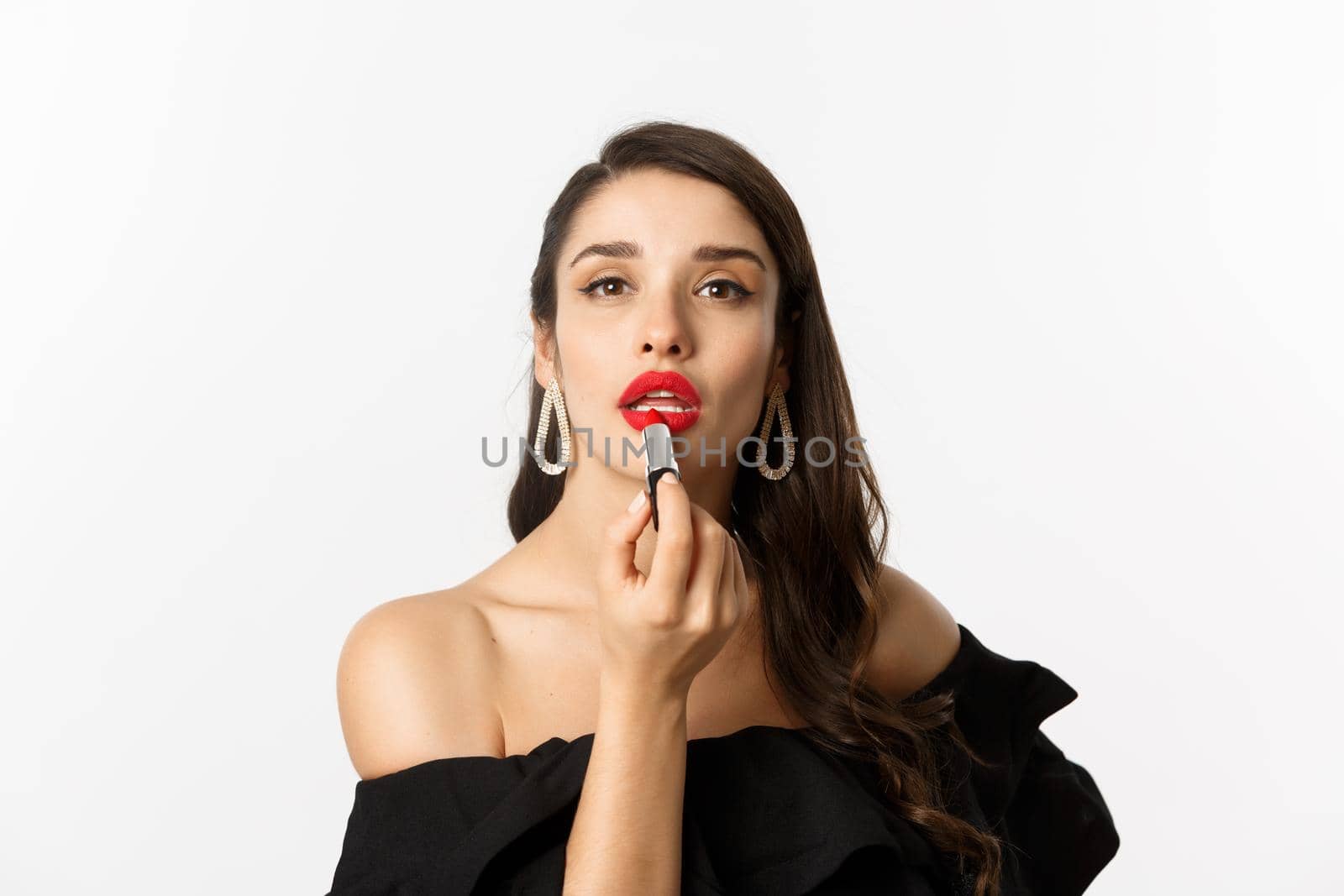 Close-up of beautiful woman applying red lipstick on lips, looking at camera like mirror, standing in black dress over white background by Benzoix