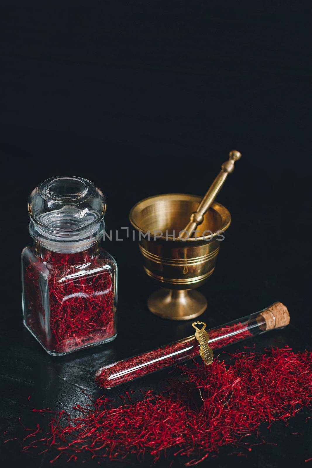 Raw organic red dried saffron spice on wooden background in vintage metal brass mortar with pestle, glass jar and tube.