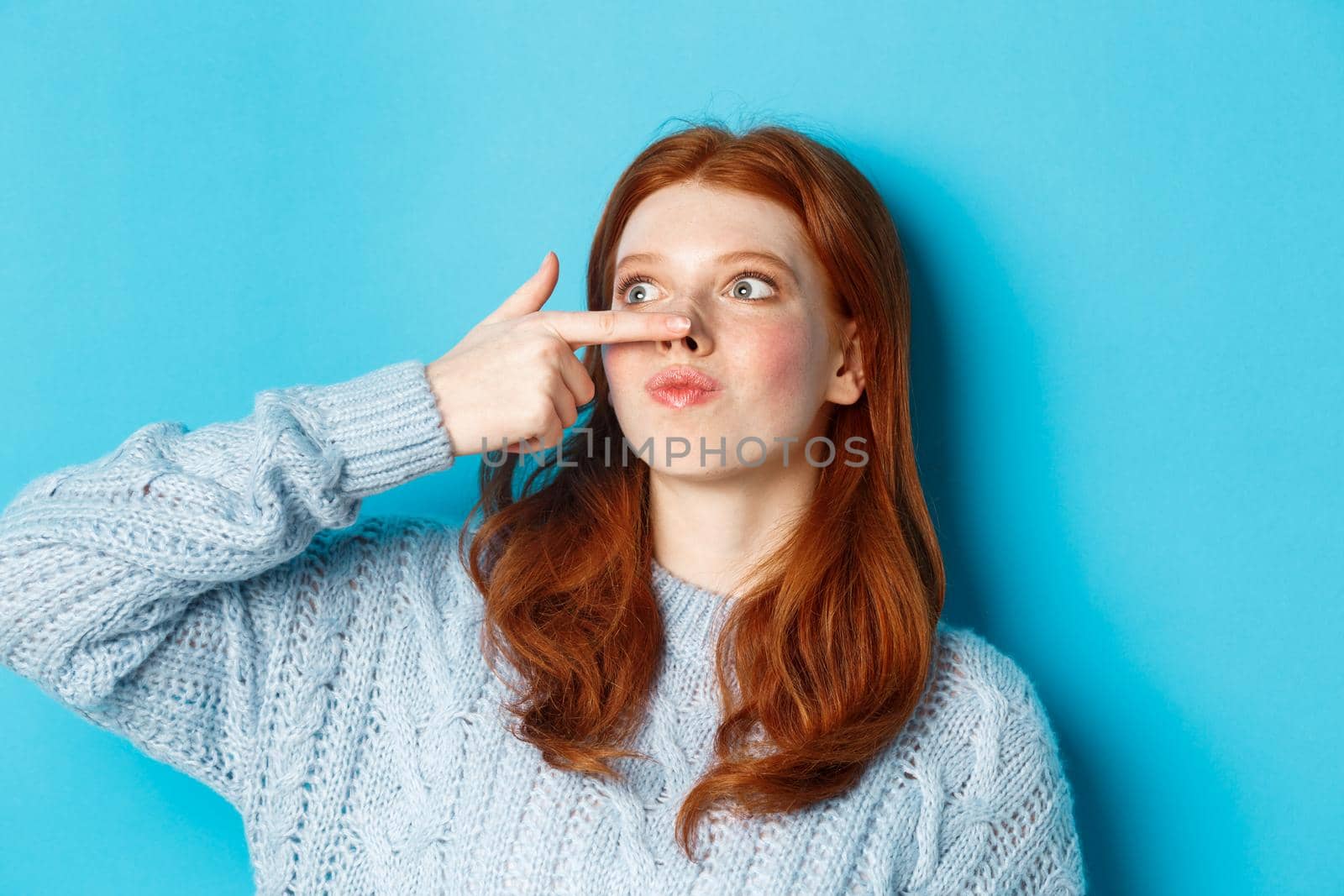 Close-up of silly redhead girl showing piggy nose, making funny grimaces and standing against blue background.