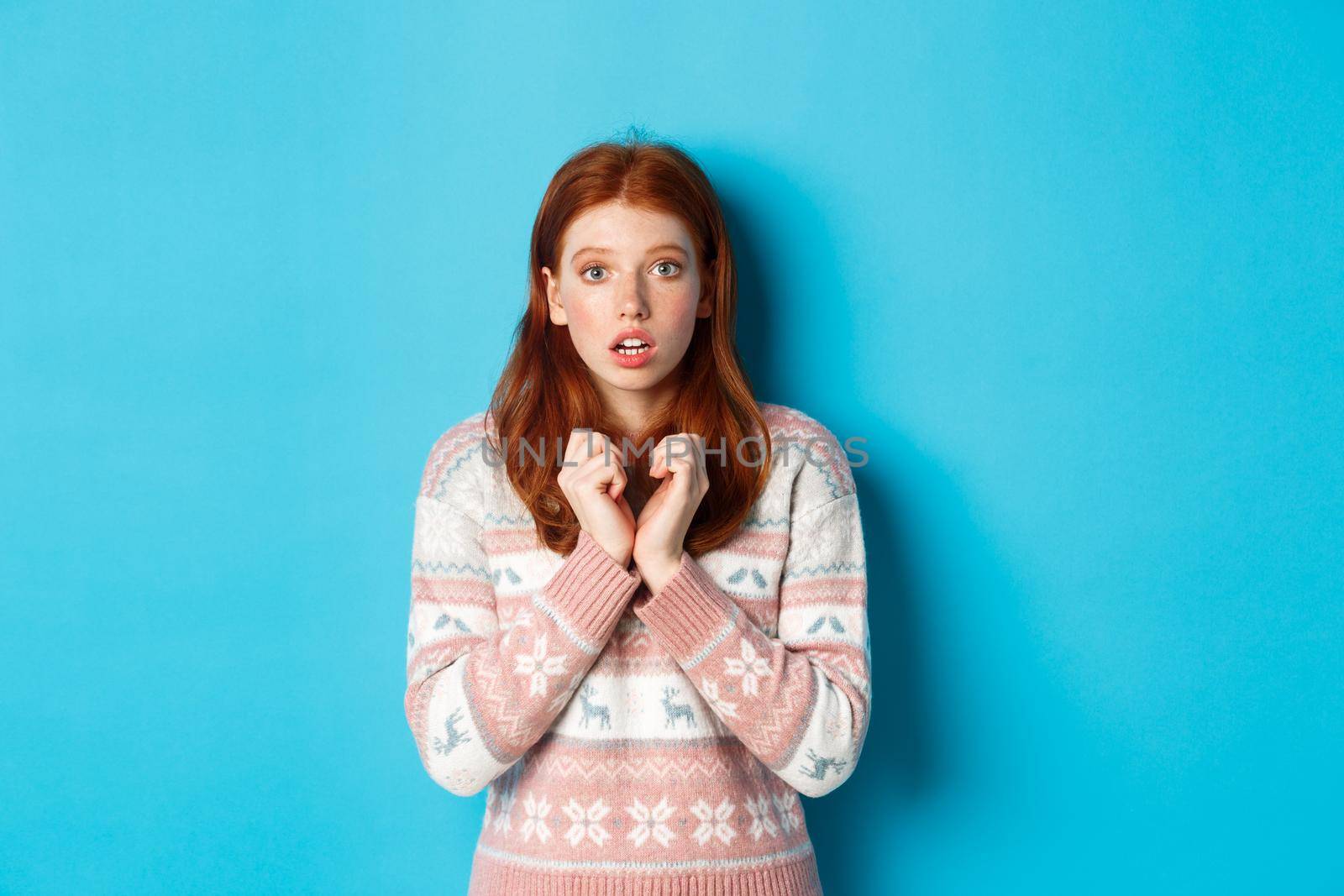Close-up of cute redhead girl looking with anticipation and worry, staring at camera, standing in winter sweater against blue background.