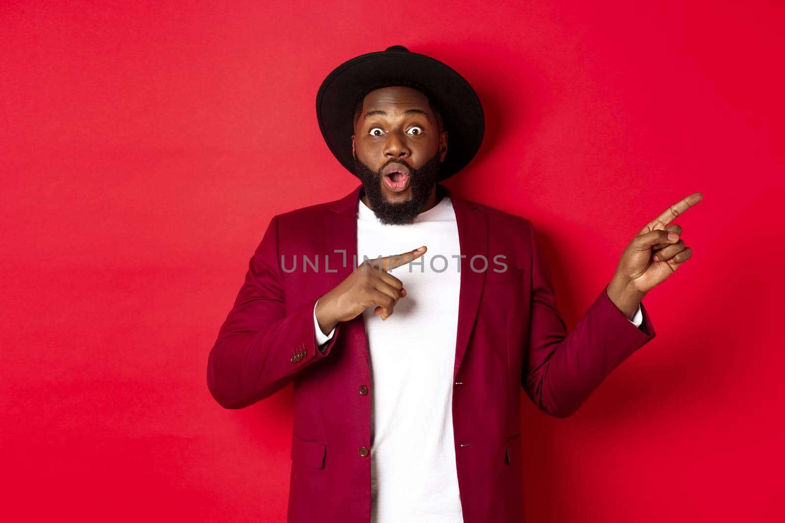 Winter holidays and shopping concept. Stylish african american man pointing fingers right at copy space for logo, standing over red background.