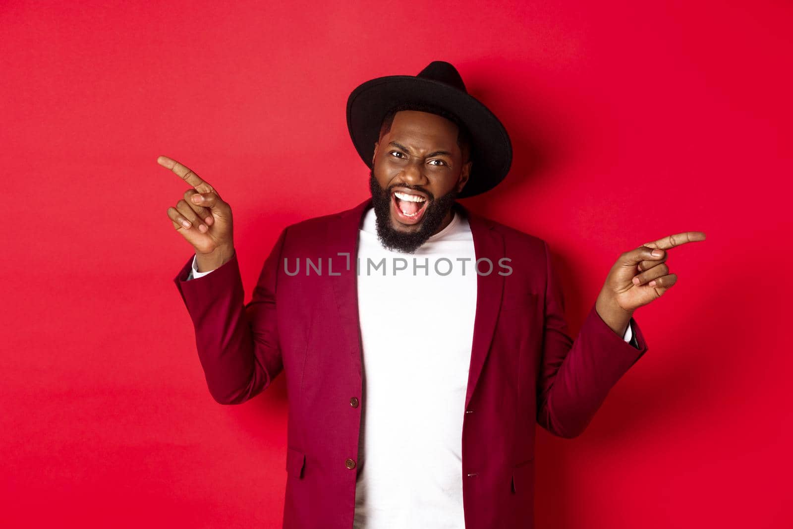 Winter holidays and shopping concept. Cheerful Black man smiling and showing two promo, pointing fingers sideways at copy spaces, red background by Benzoix