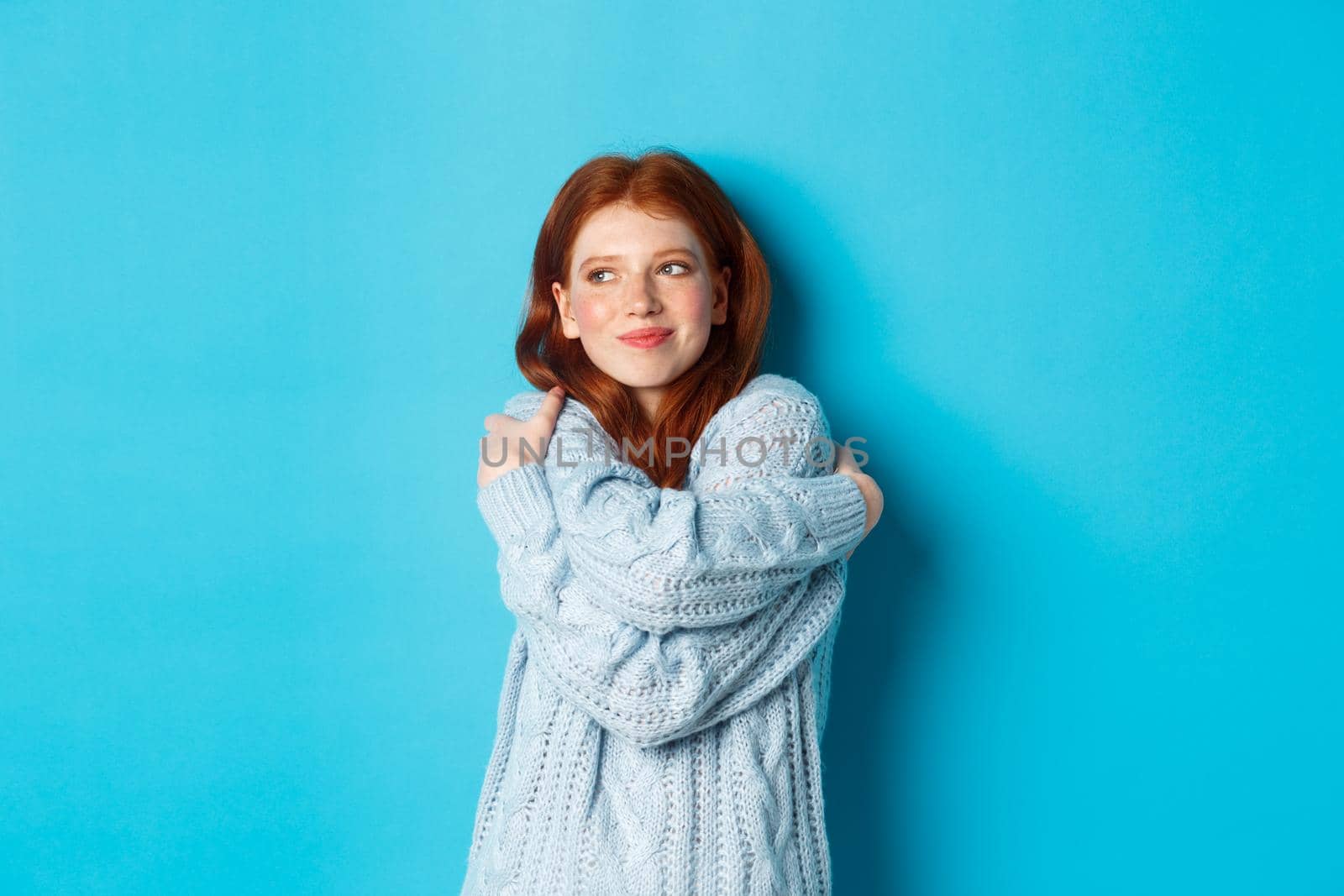 Happy cute redhead girl hugging herself, wearing comfortable and warm sweater, smiling at camera, standing over blue background by Benzoix