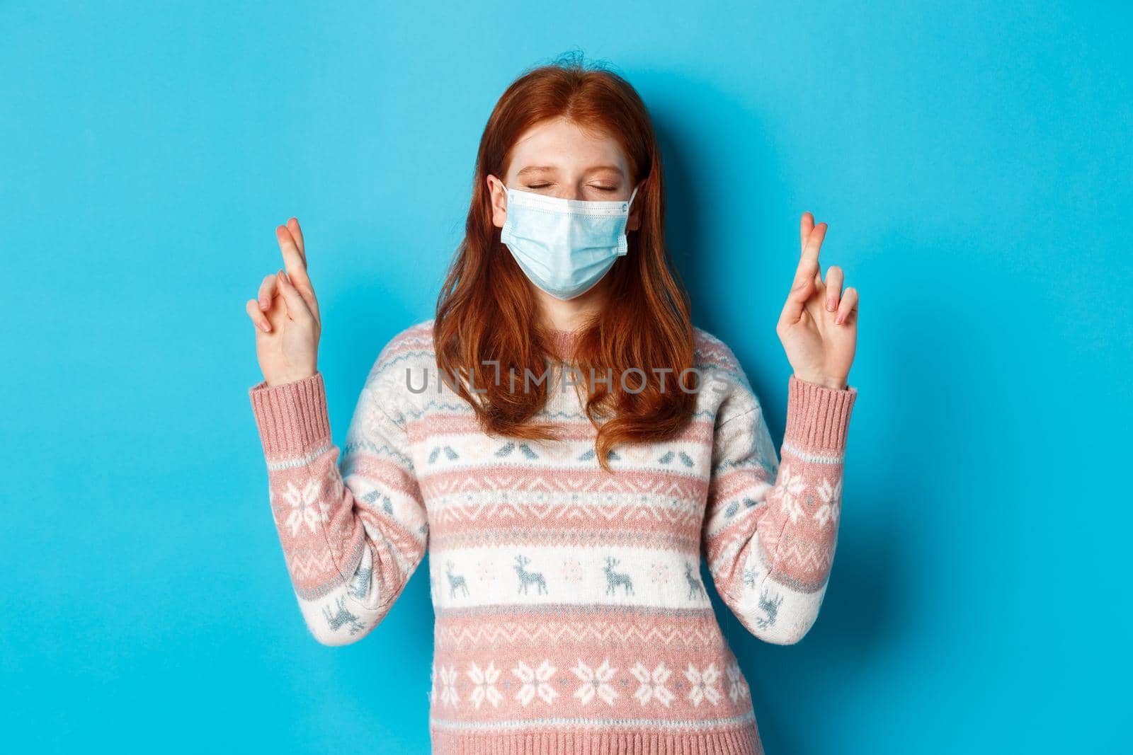Winter, coronavirus and social distancing concept. Cute hopeful girl with red hair, wearing face mask, cross fingers and making wish, standing over blue background.