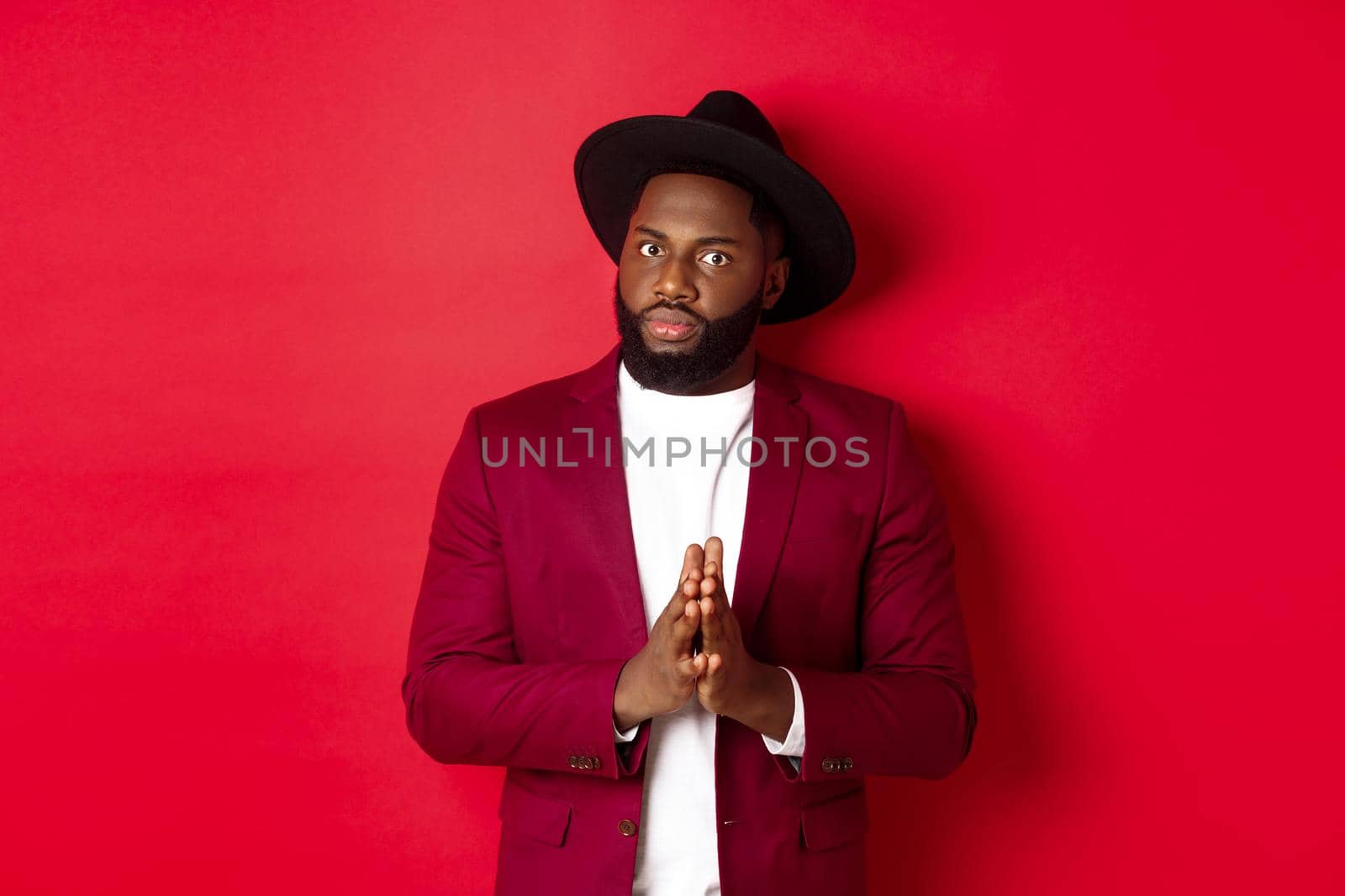 Serious looking man getting ready to business, holding hands pressed together and nervously staring at camera, listening closely, standing against red background by Benzoix