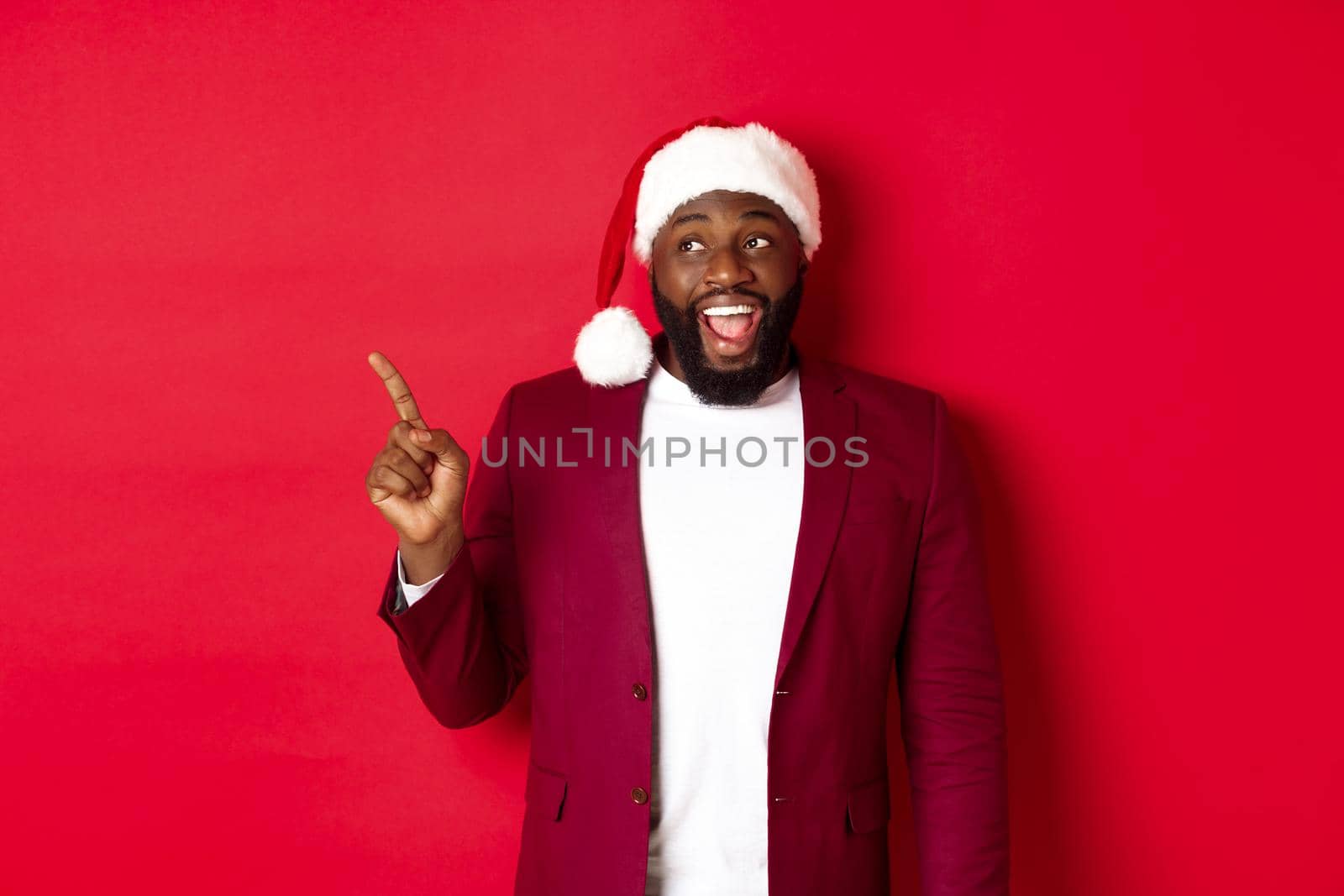 Christmas, party and holidays concept. Cheerful Black guy in santa hat pointing fingers left, showing banner, standing amused against red background by Benzoix