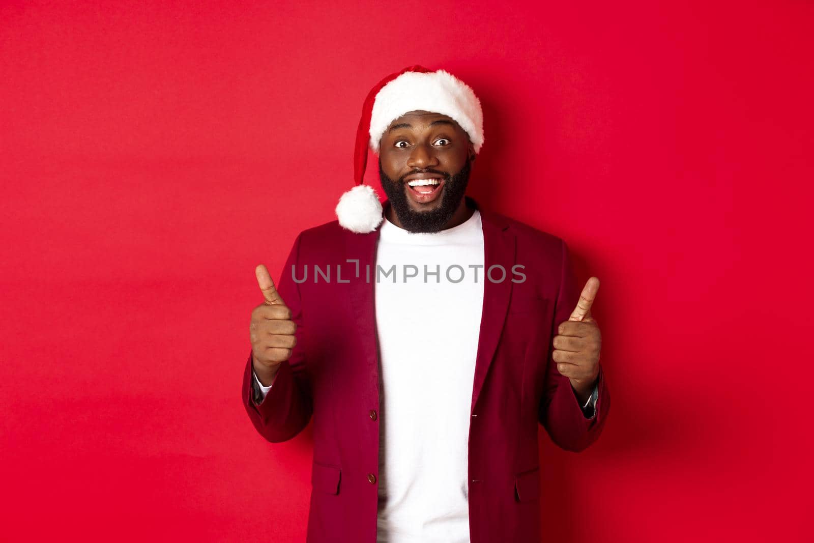 Christmas, party and holidays concept. Cheerful Black man in santa hat showing thumbs-up, celebrating New Year, approve and agree, standing against red background.