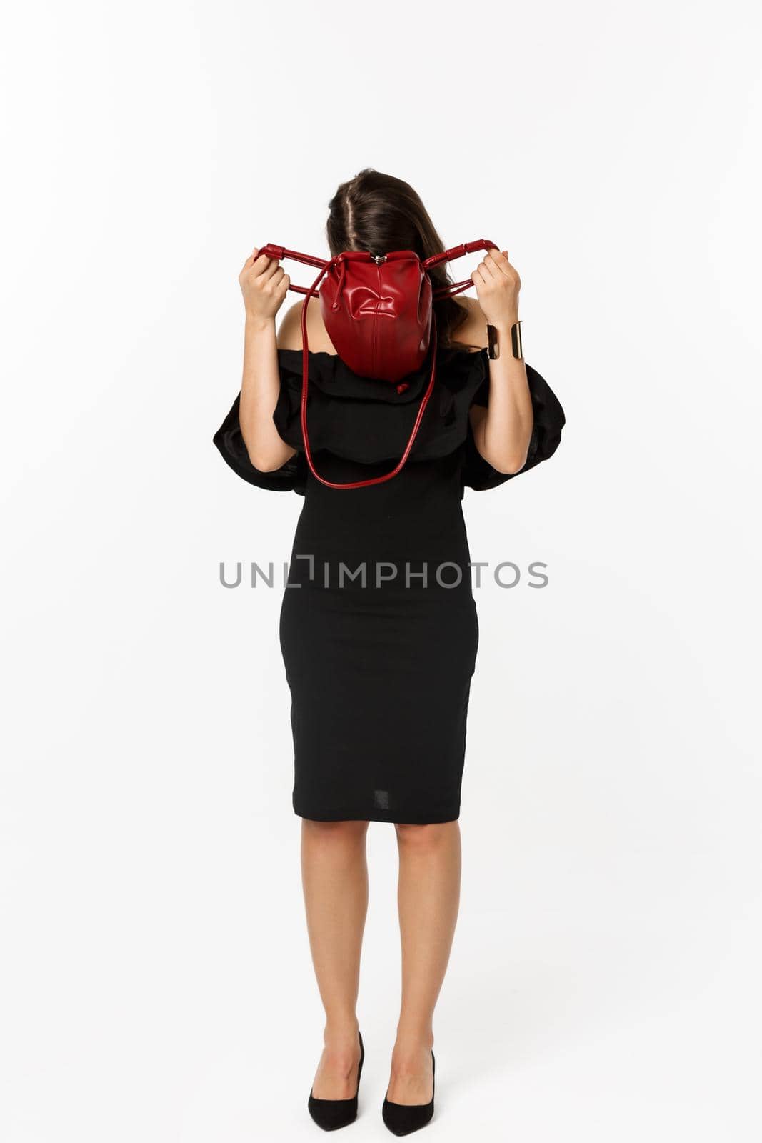 Beauty and fashion concept. Full length of young woman sticking head inside purse and searching something, wearing black dress and high heels, standing over white background.