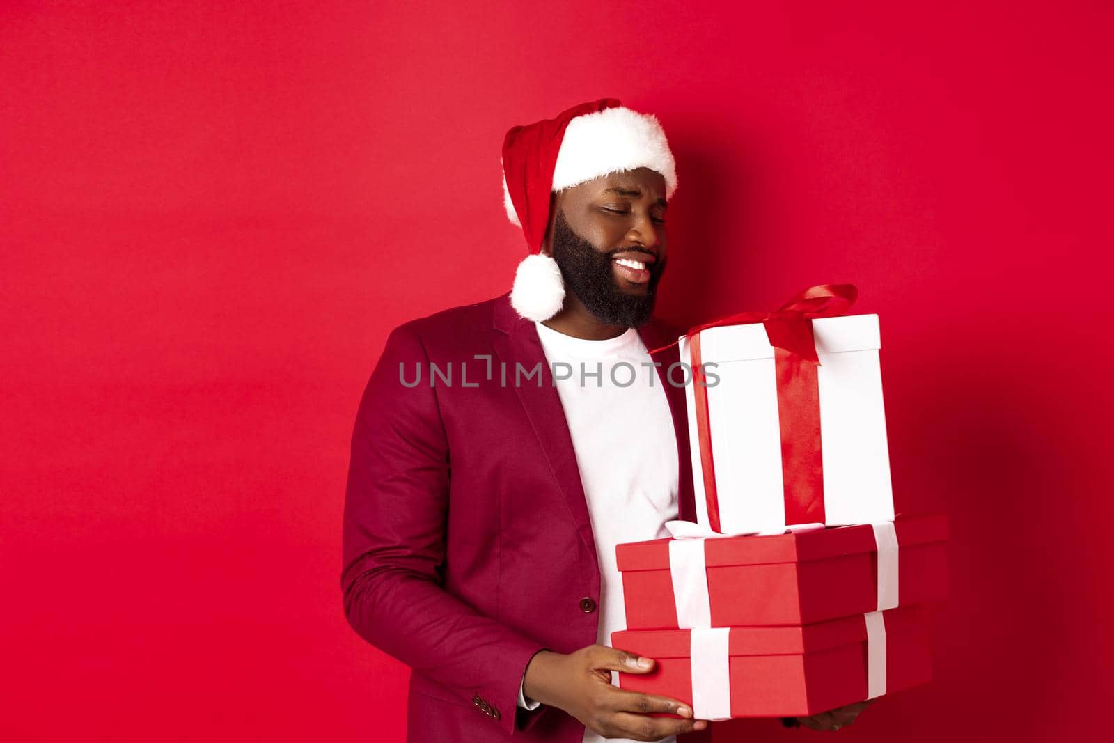 Christmas, New Year and shopping concept. Happy african amaerican man looking touched and thankful at xmas presents, receiving gifts, wearing santa hat, red background.
