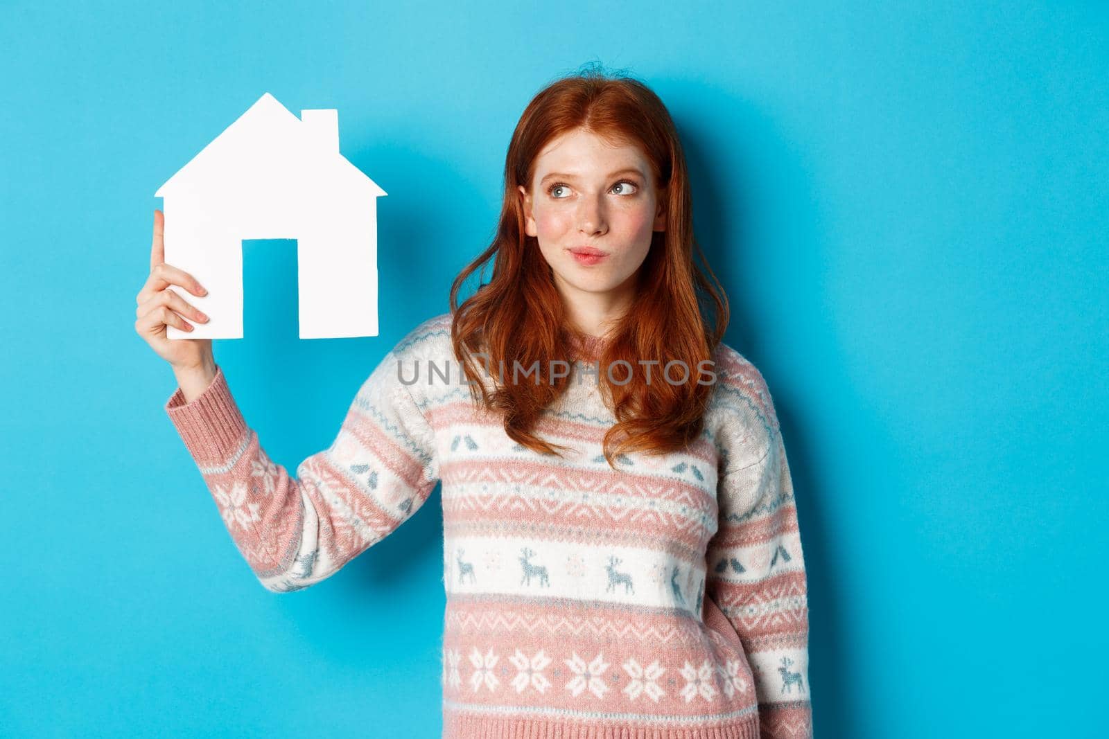 Real estate concept. Image of thoughtful redhead girl showing paper house model and thinking, searching for home or flat, standing against blue background.