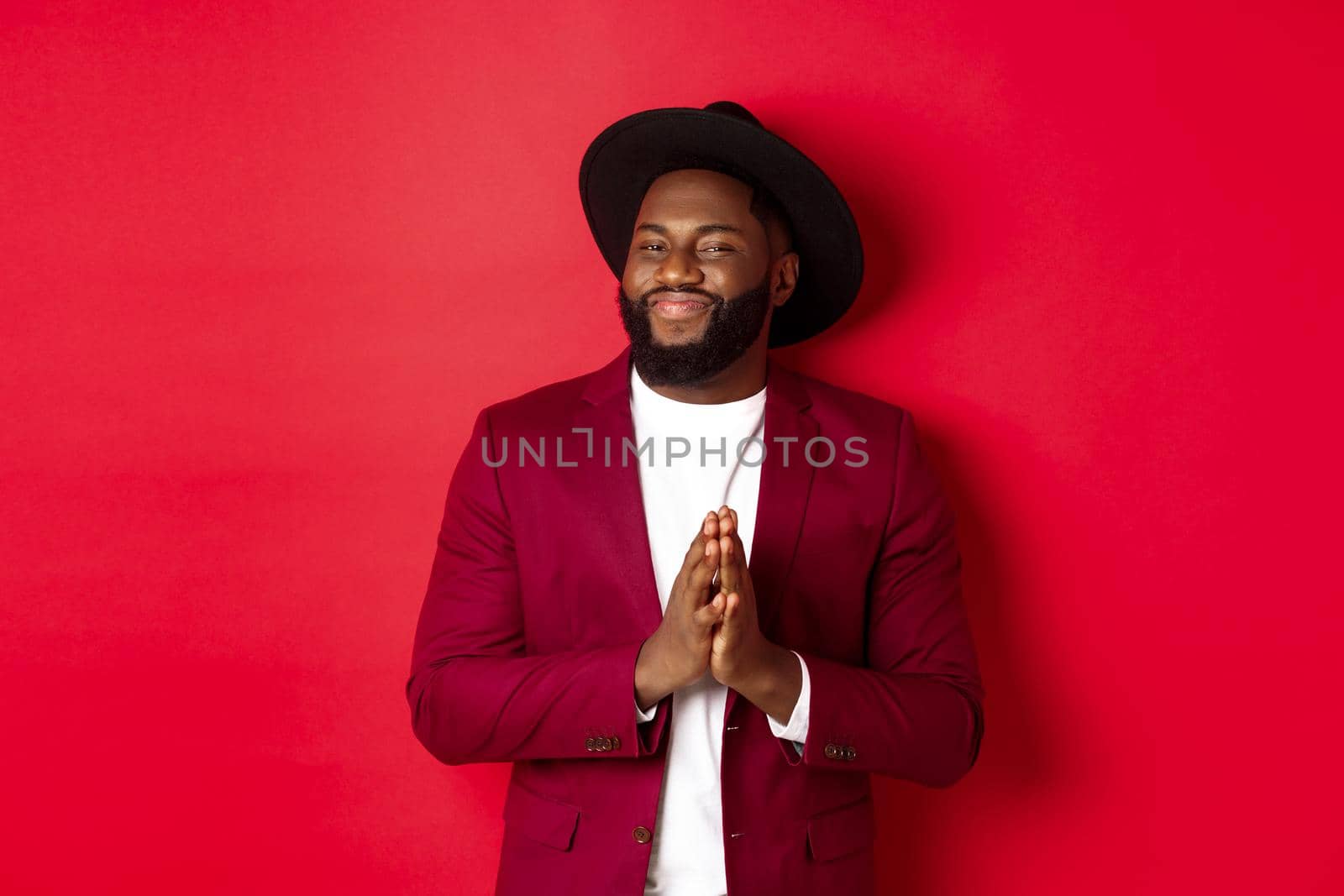Handsome successful Black man relish profit, rub hands and smiling satisfied, standing against red background in party outfit by Benzoix