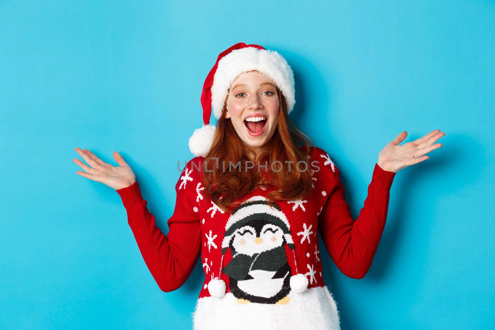 Happy holidays concept. Cheerful redhead girl in xmas sweater and santa hat, raising hands up and wishing merry Christmas, standing against blue background.