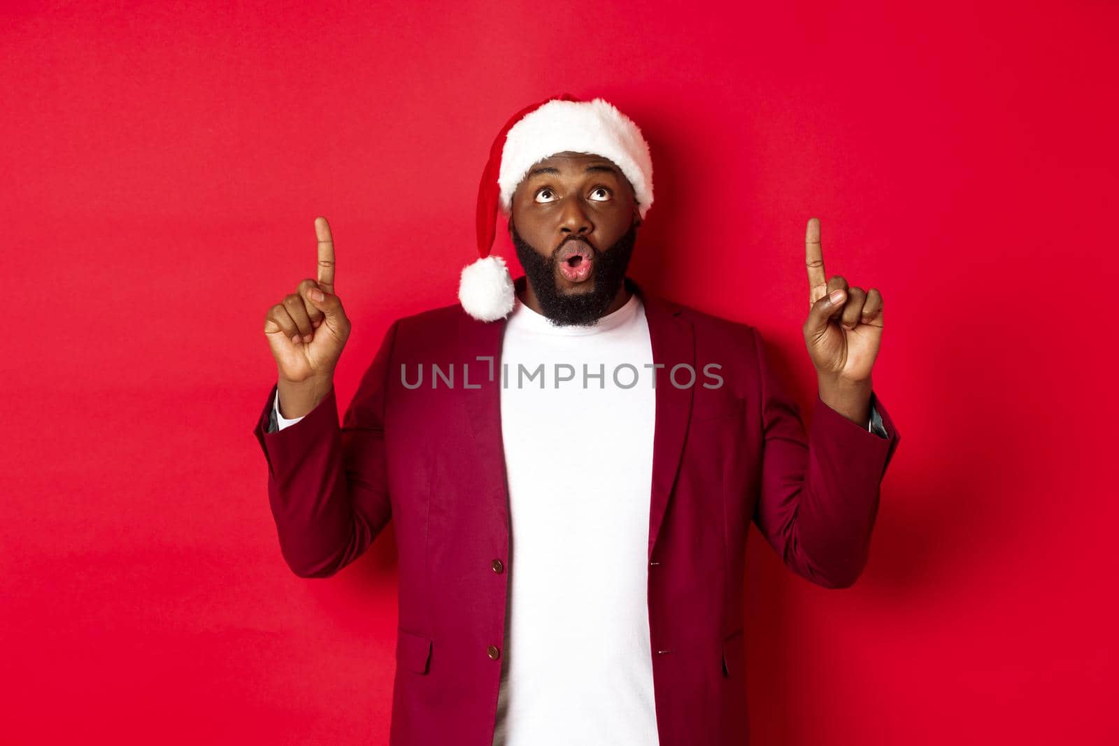 Christmas, party and holidays concept. Impressed Black man in santa hat pointing fingers up, saying wow and staring at camera, checking out promo offer, red background by Benzoix