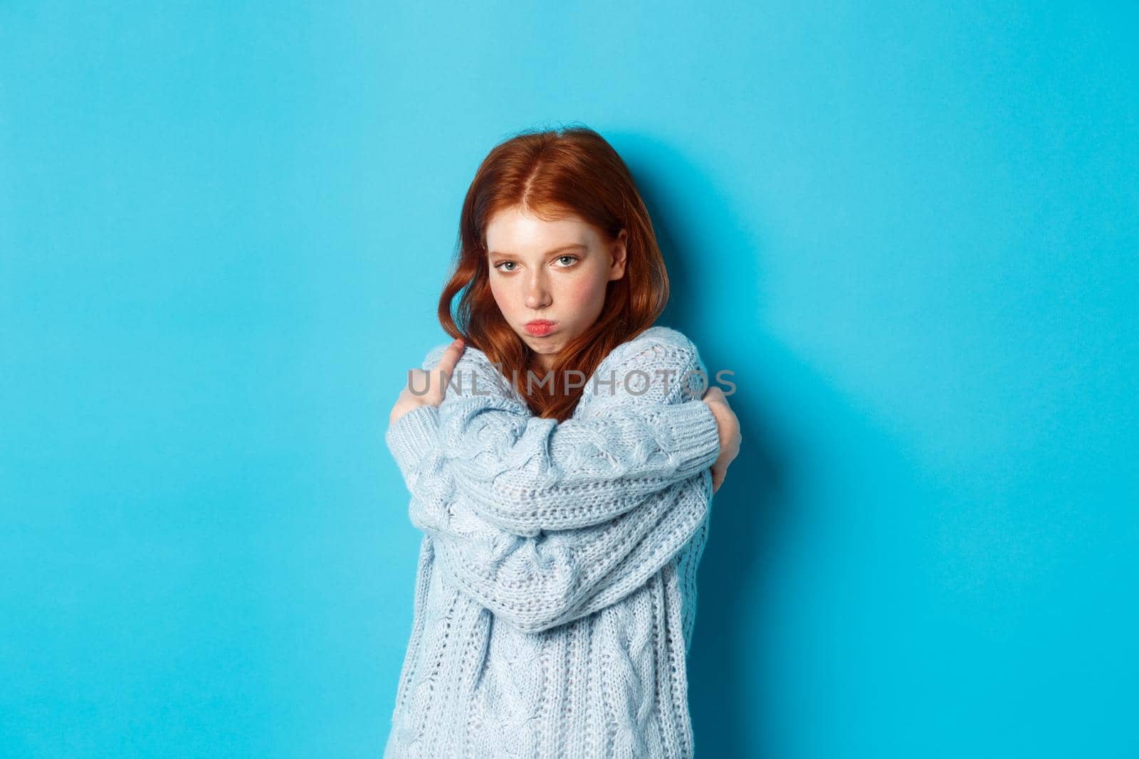 Silly and cute redhead girl pucker lips and looking offended, comforting herself by cuddling, embracing body and staring at camera defensive, blue background.