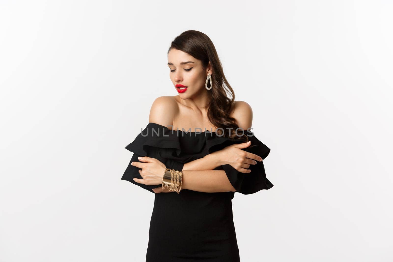 Beauty and fashion concept. Sensual and attractive woman in black dress and red lips, hugging herself and gently looking down, standing over white background.