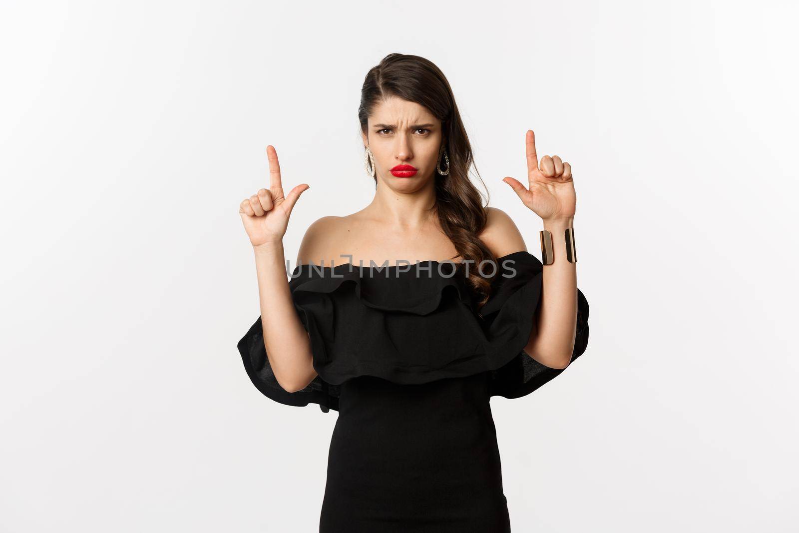 Fashion and beauty. Disappointed woman sulking upset, pointing fingers up and complaining, standing dissatisfied in black dress, white background.