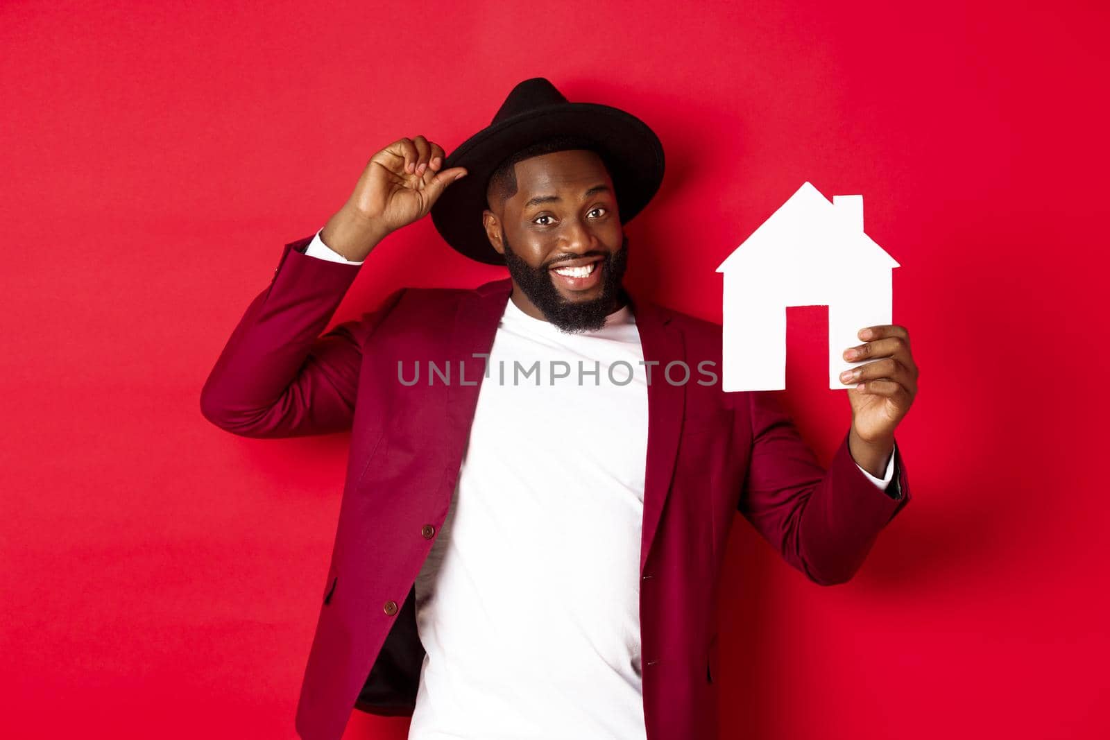 Real estate. Cheerful Black man showing paper house and smiling, recommending broker, standing over red background.