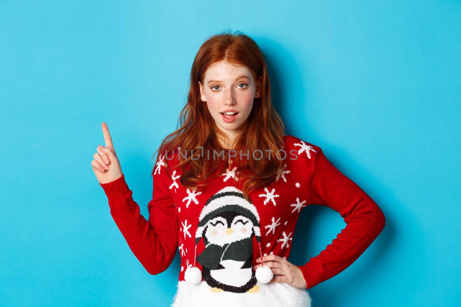 Winter holidays and Christmas Eve concept. Sassy redhead girl in xmas sweater, pointing upper left corner and staring at camera, hinting on promo offer, blue background.