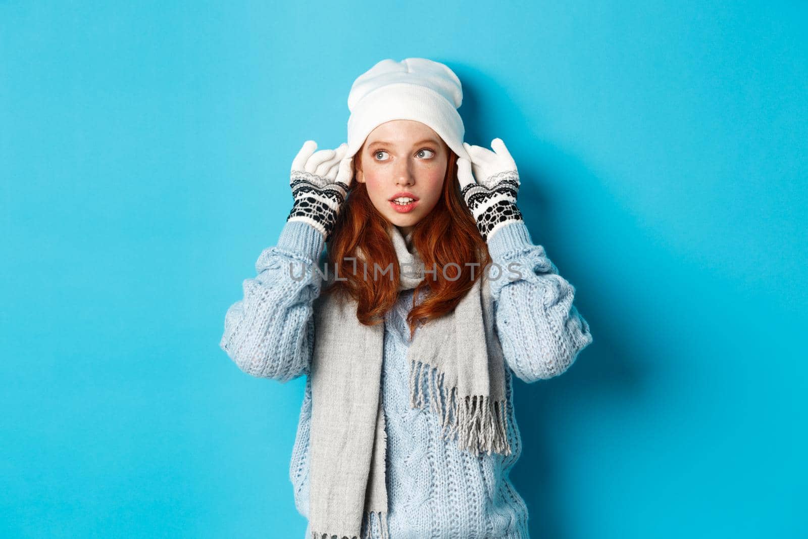 Winter and holidays concept. Cute redhead girl going outside, put on beanie and gloves, looking left, standing over blue background.