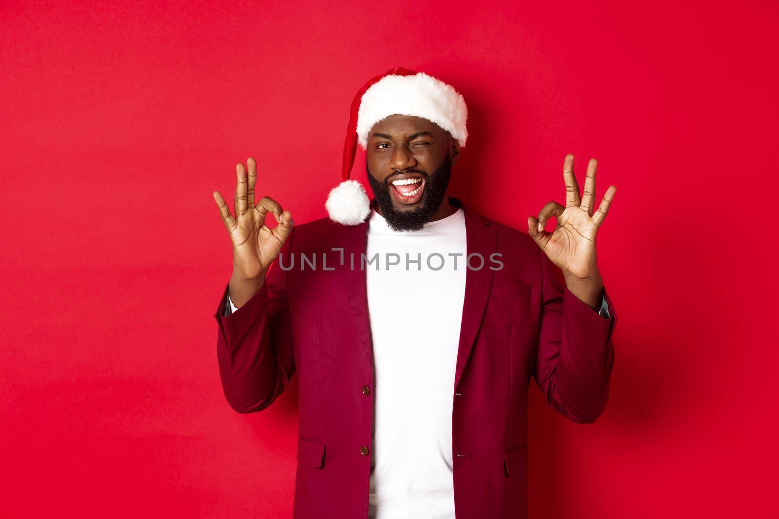 Christmas, party and holidays concept. Handsome african american man in santa hat, winking and showing ok signs, standing against red background.
