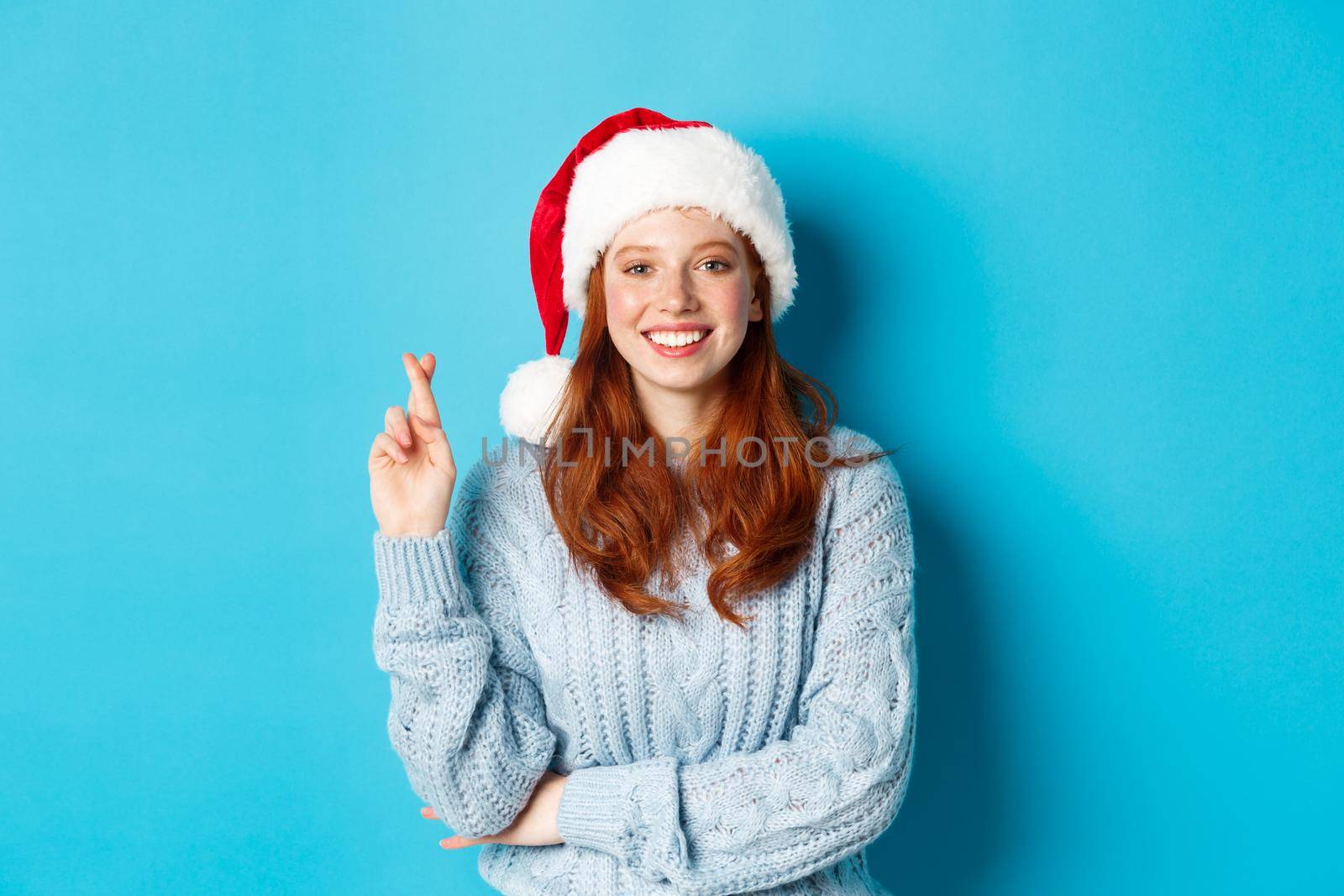 Winter holidays and Christmas Eve concept. Hopeful redhead girl in Santa hat, making wish on xmas, cross fingers for good luck and smiling, standing over blue background by Benzoix
