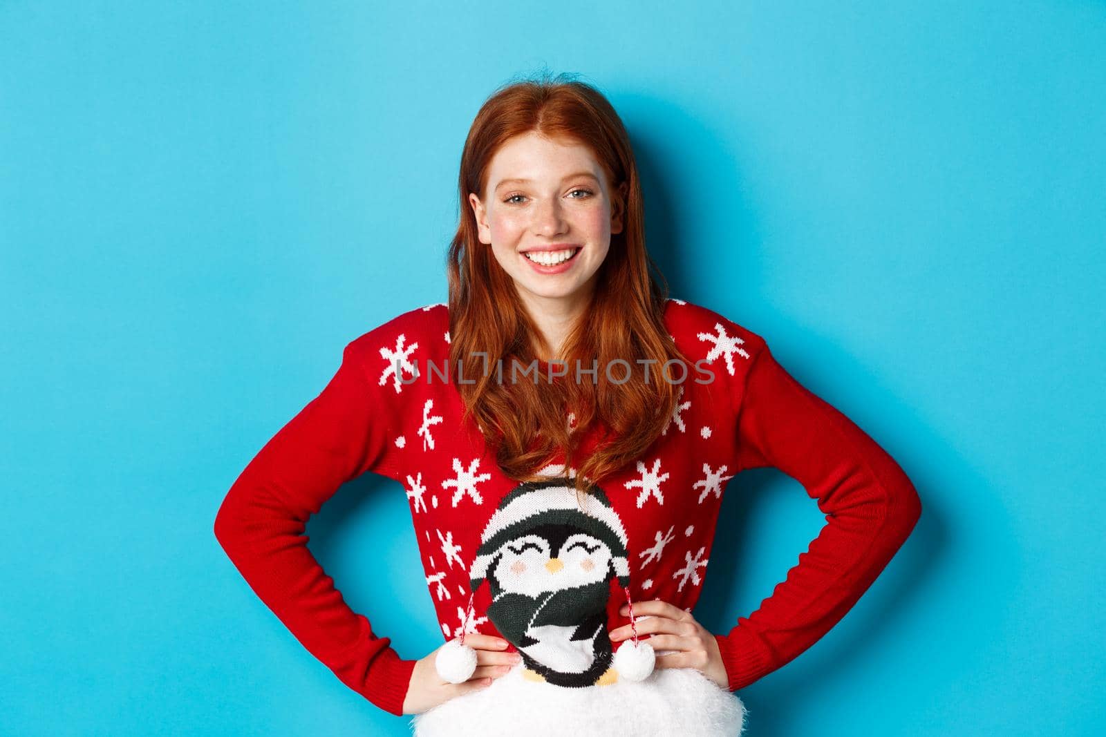 Winter holidays and Christmas Eve concept. Beautiful teenage redhead girl in xmas sweater looking at camera, smiling carefree, holding hands on waist, blue background.