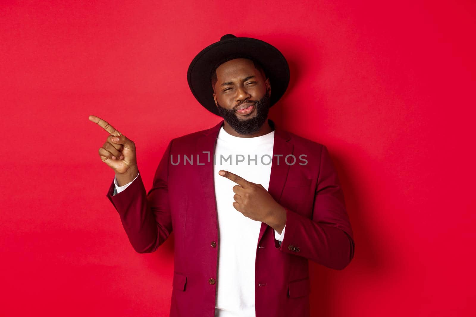 Winter holidays and shopping concept. Skeptical african american man looking unsure, pointing left at logo, having doubts, standing against red background.