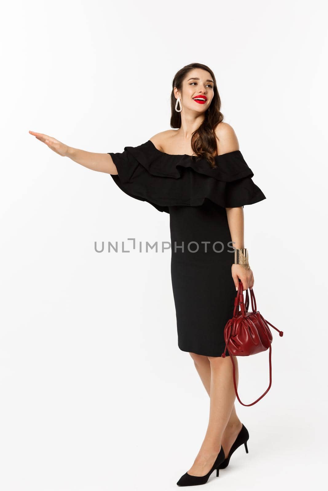 Beauty and fashion concept. Full length of glamour woman in black dress and high heels raising hand to stop taxi, need a ride, standing over white background by Benzoix