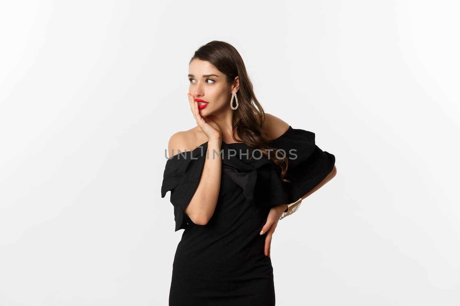 Fashion and beauty. Image of stylish beautiful woman in black dress and makeup, looking left with temptation, touching red lips, standing over white background by Benzoix