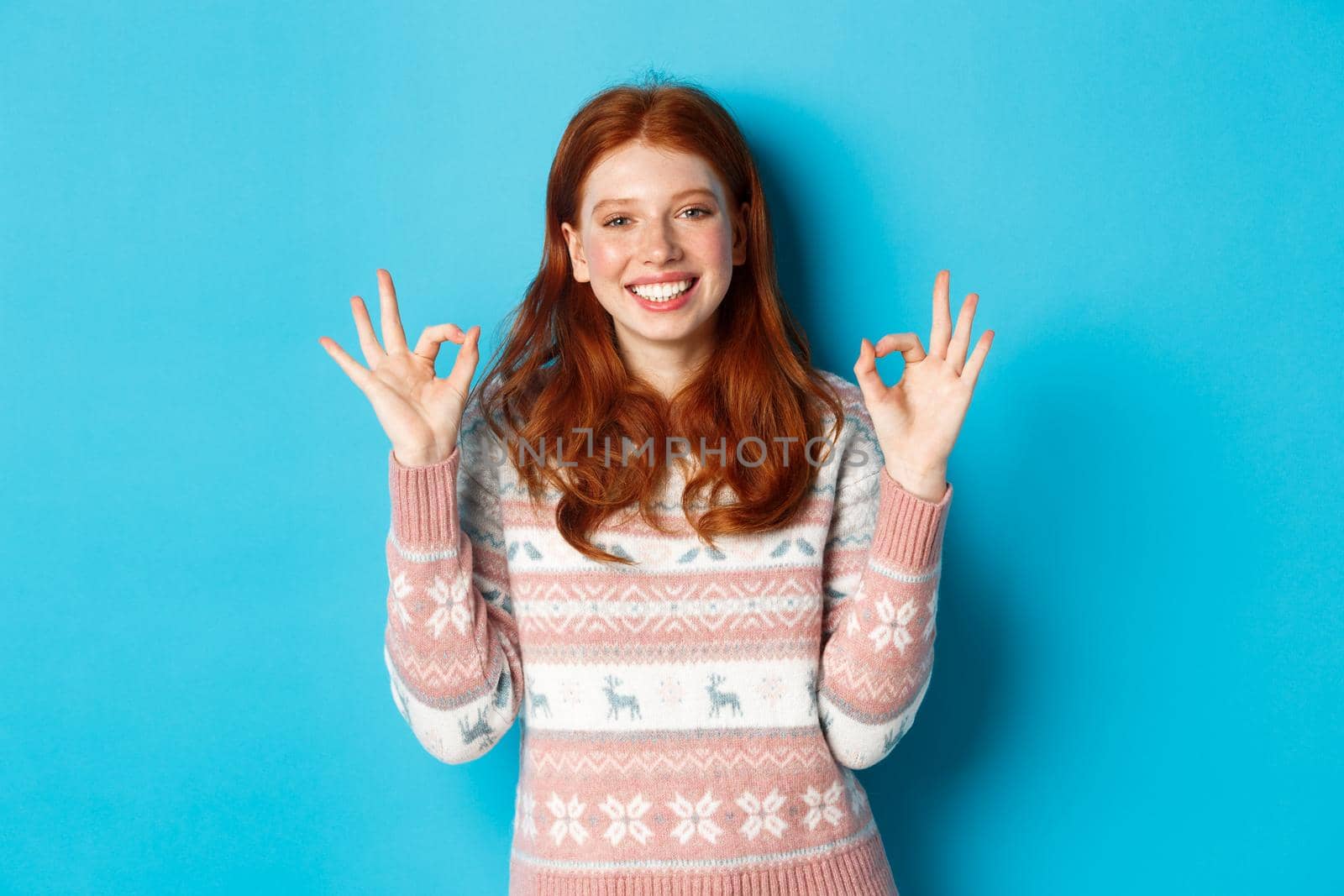 Cute redhead girl in sweater showing okay signs, smiling pleased, approve and like, praise excellent choice, standing over blue background by Benzoix