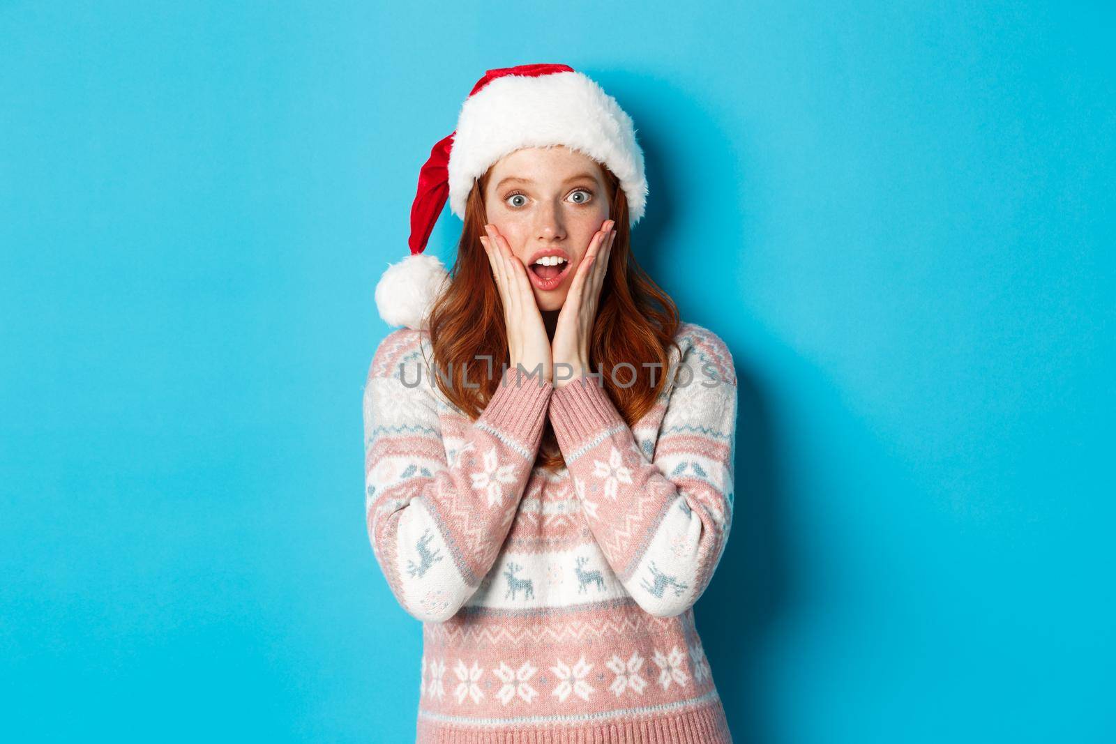 Winter and Christmas Eve concept. Impressed redhead girl in santa hat hear amazing news, touching cheeks and open mouth fascinated, standing against blue background.