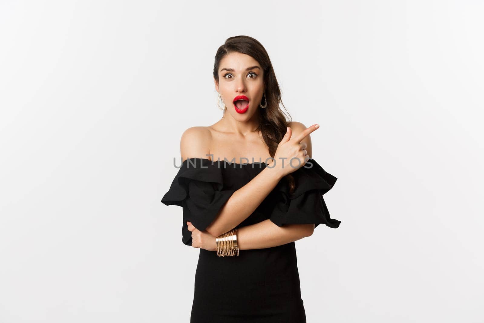 Fashion and beauty. Beautiful woman in black dress, red lips, pointing finger right at promo offer, looking amazed at camera, white background.
