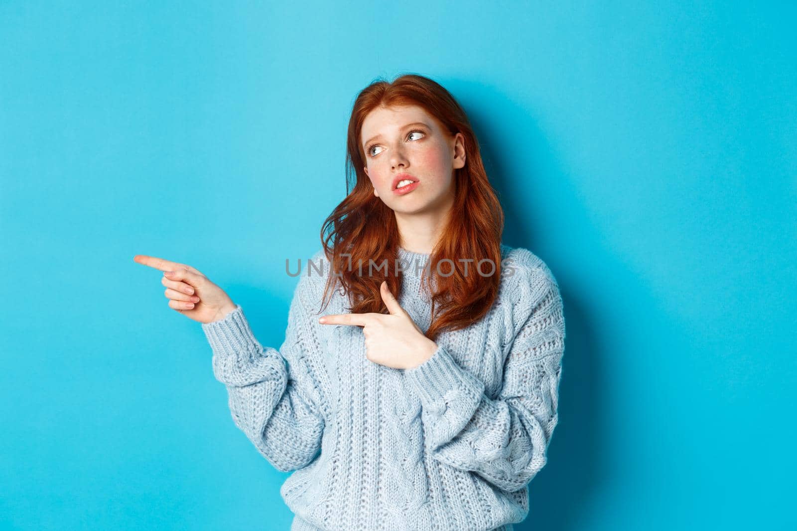 Annoyed teenage redhead girl roll eyes, pointing fingers left at something boring or lame, standing irritated over blue background.