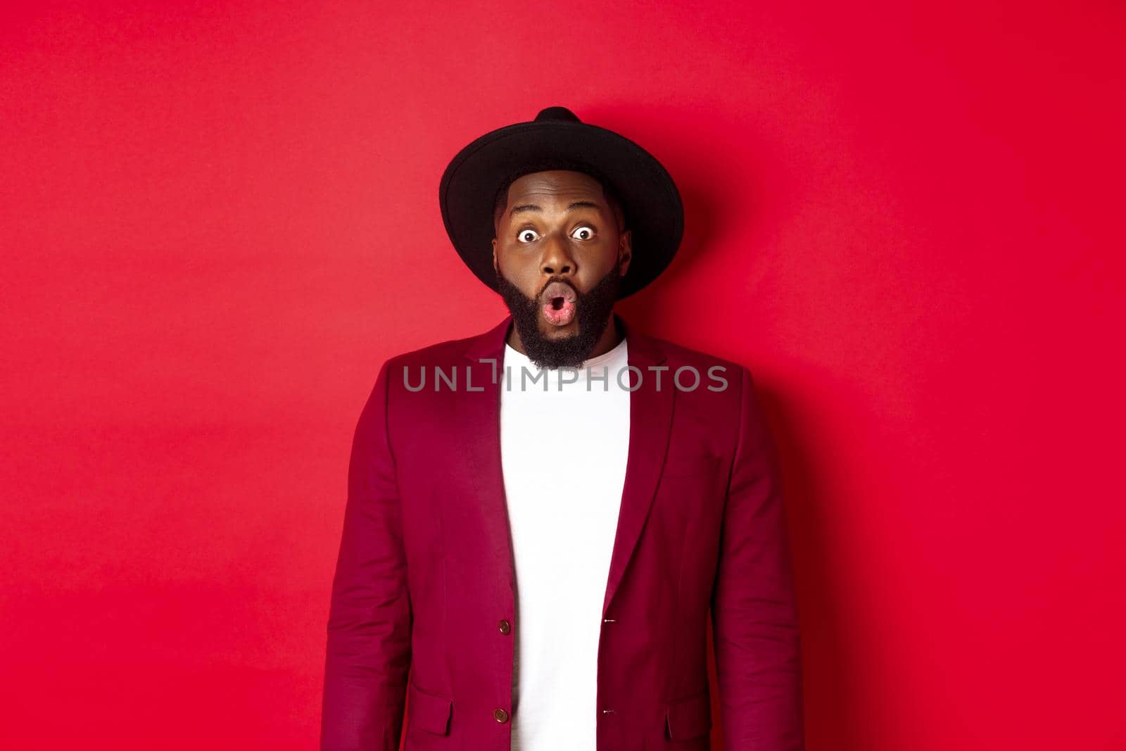 Fashion and party concept. Impressed Black man in classy outfit staring with complete disbelief at camera, gasping and looking surprised, standing over red background.