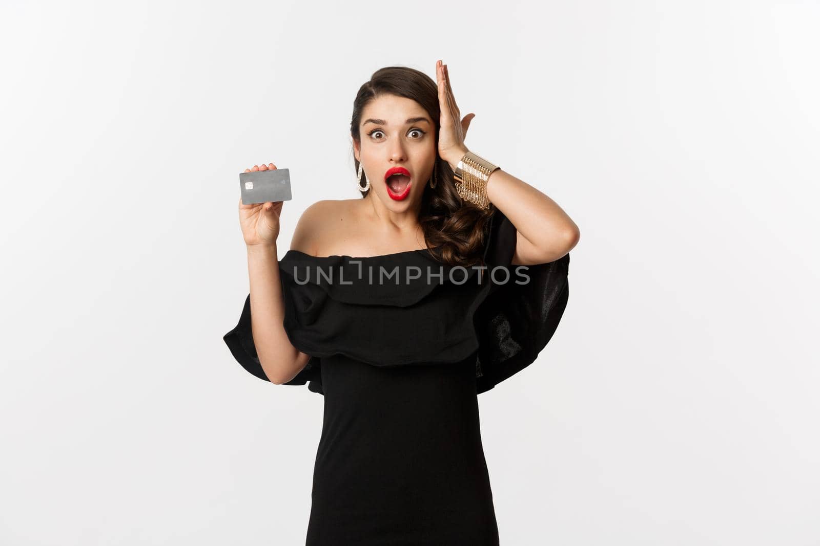 Fashion and shopping concept. Shocked woman in black dress showing credit card, looking in awe at camera, white background.