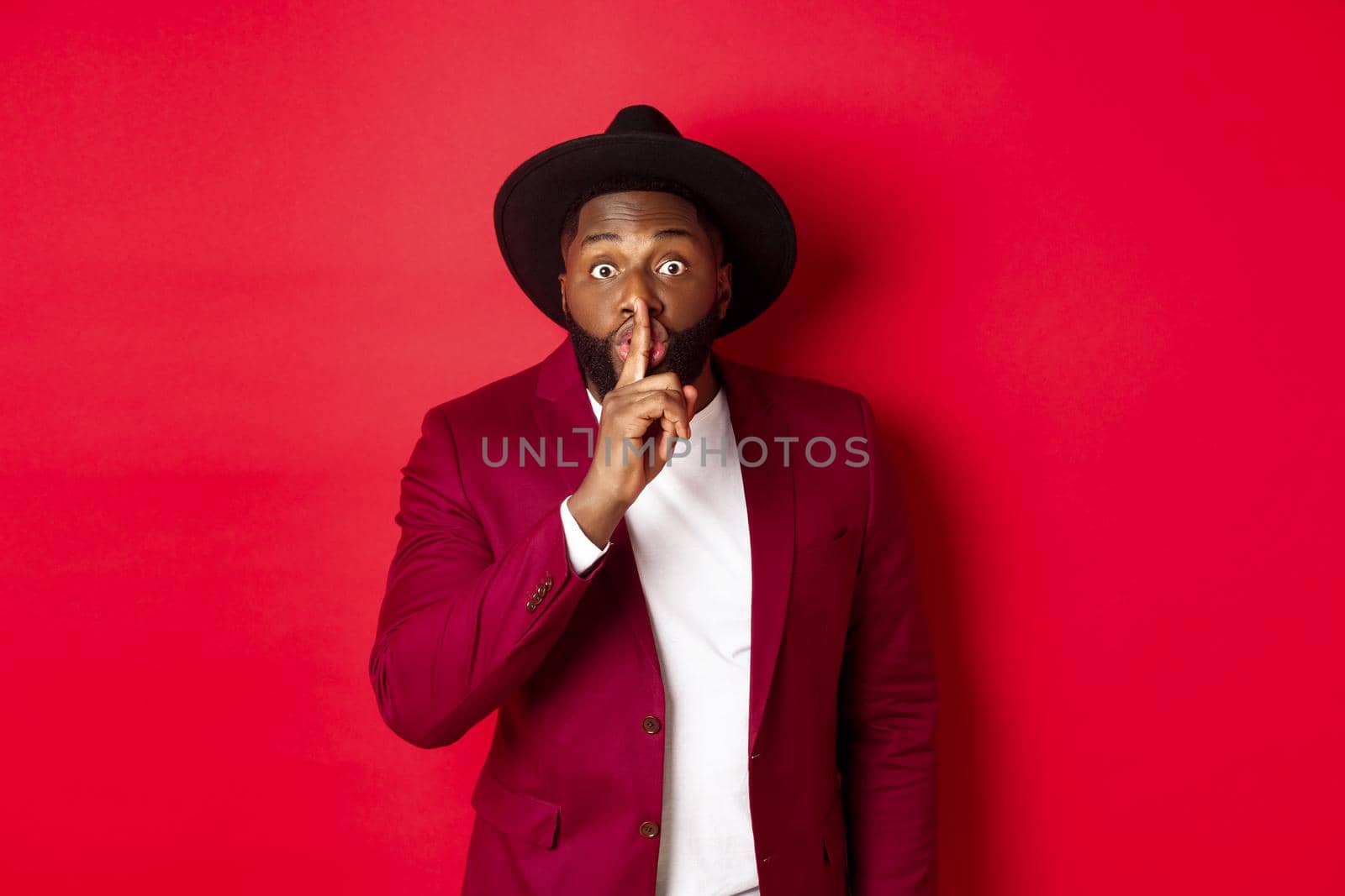 Fashion and party concept. African american man in classy hat hushing, asking keep secret, preparing surprise, standing over red background by Benzoix