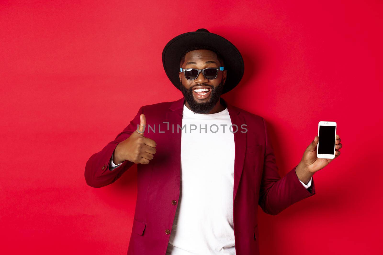 Handsome and stylish Black man showing phone screen and thumb up at camera, recommending online store app, red background.