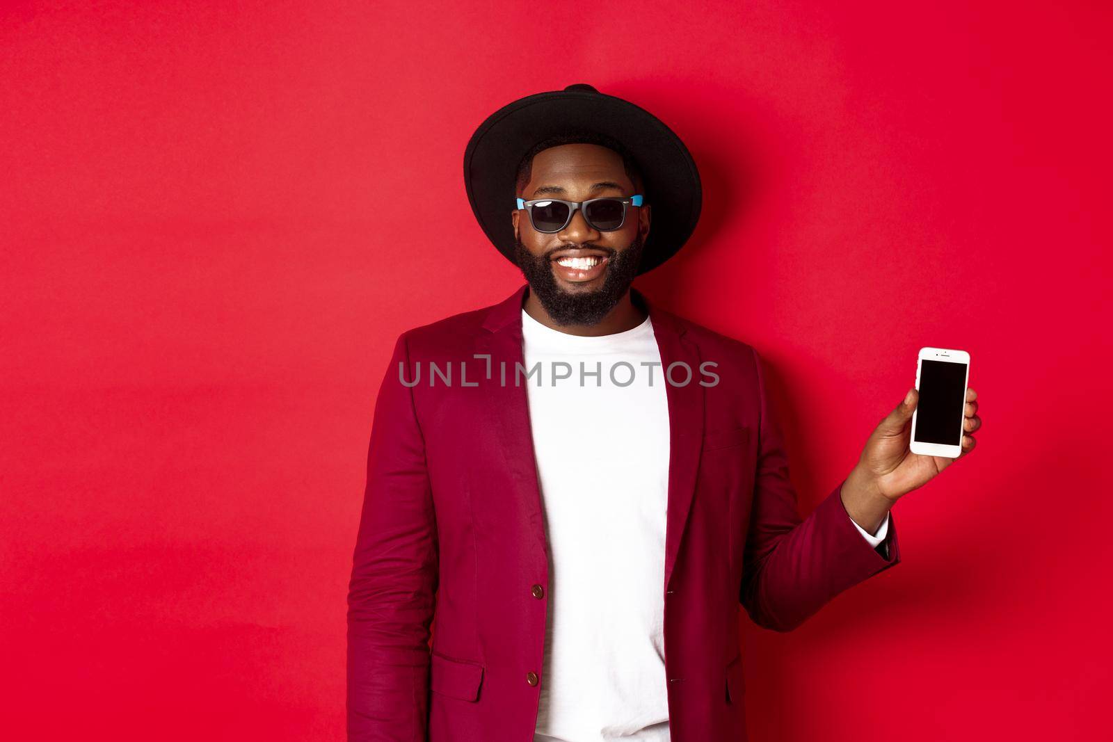 Handsome and stylish Black man showing phone screen at camera, recommending online store or application, standing over red background.