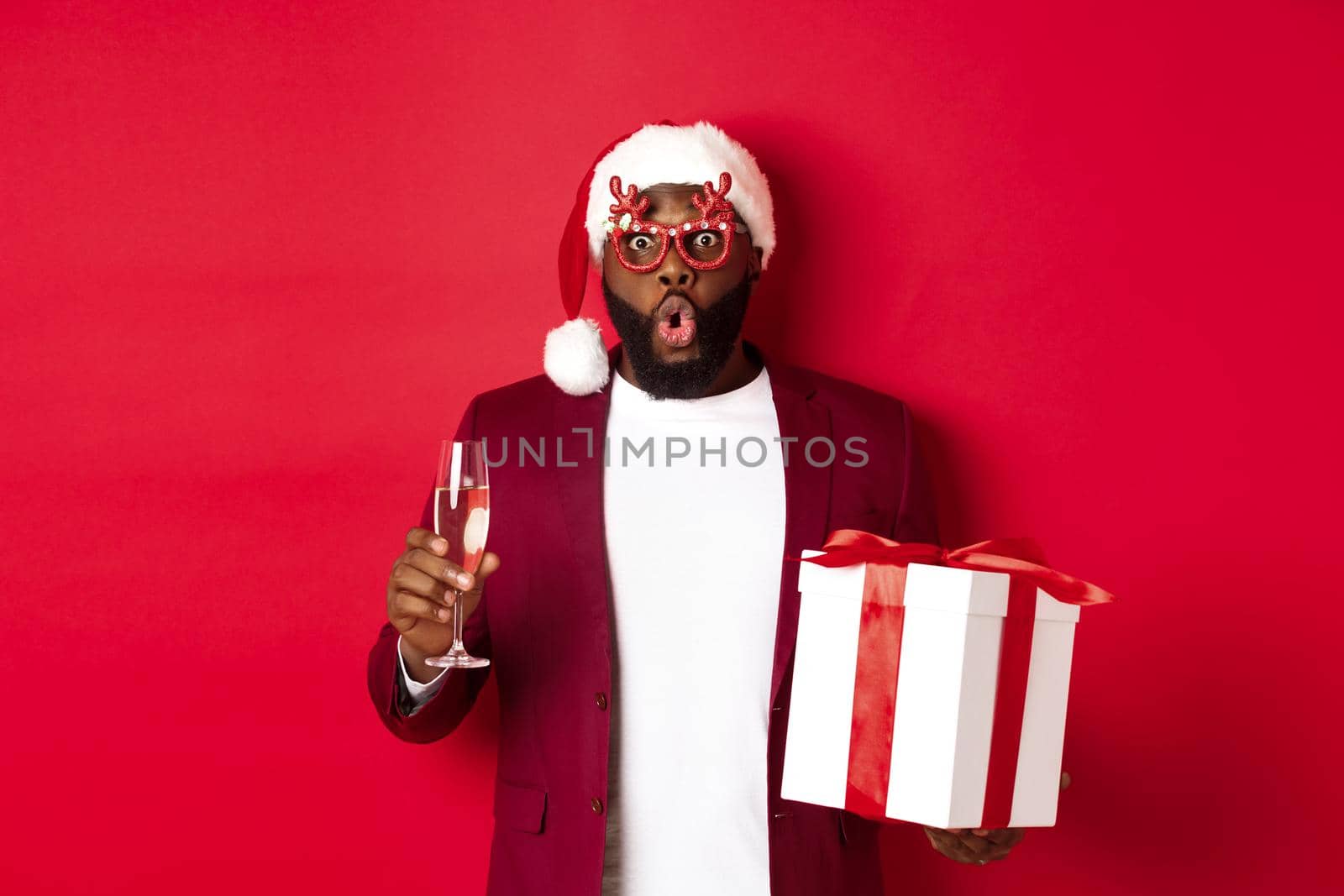 Christmas. Handsome african american man in party glasses and santa hat, holding new year gift and glass of champagne, wishing happy holidays, red background.