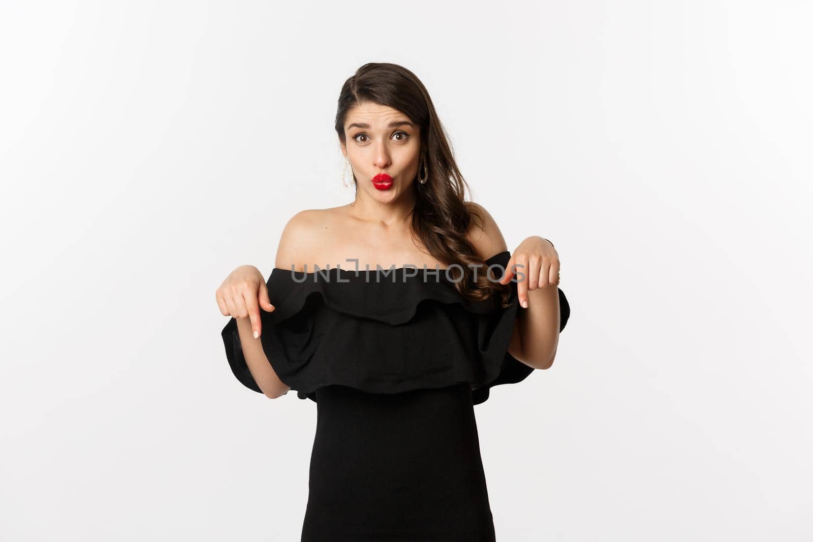 Fashion and beauty. Elegant woman in black dress pointing fingers down, showing promo and smiling, standing over white background.