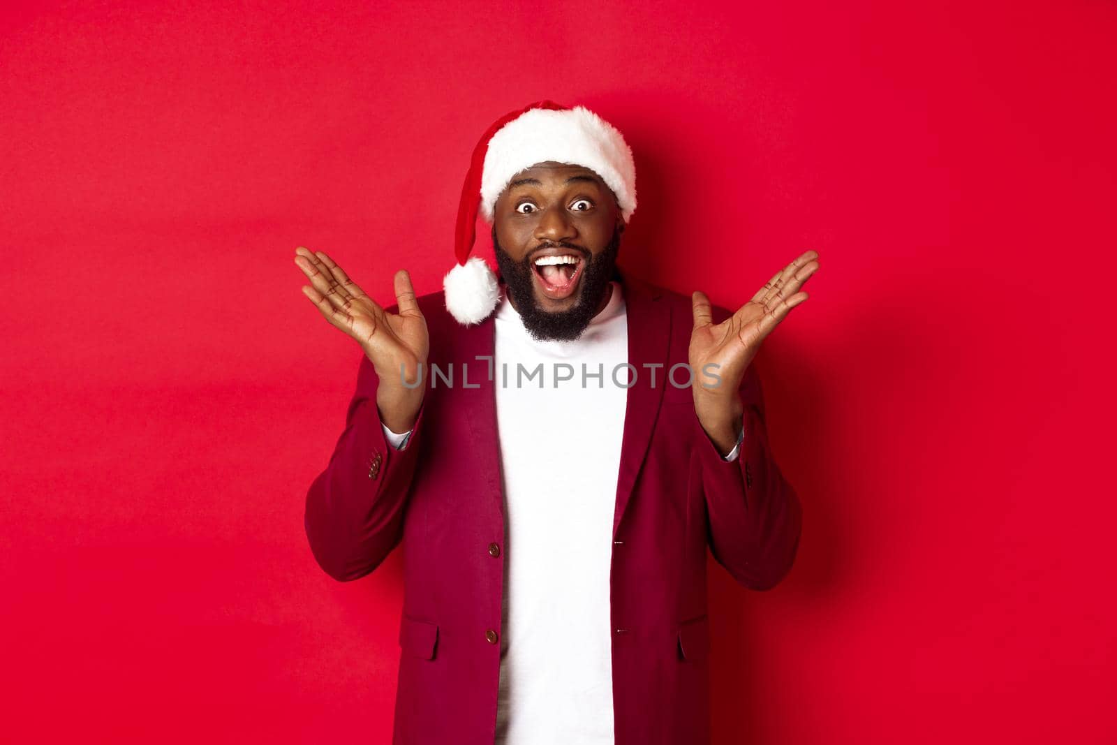 Christmas, party and holidays concept. Surprised and happy african american man in santa hat, looking with rejoice at camera, standing against red background.