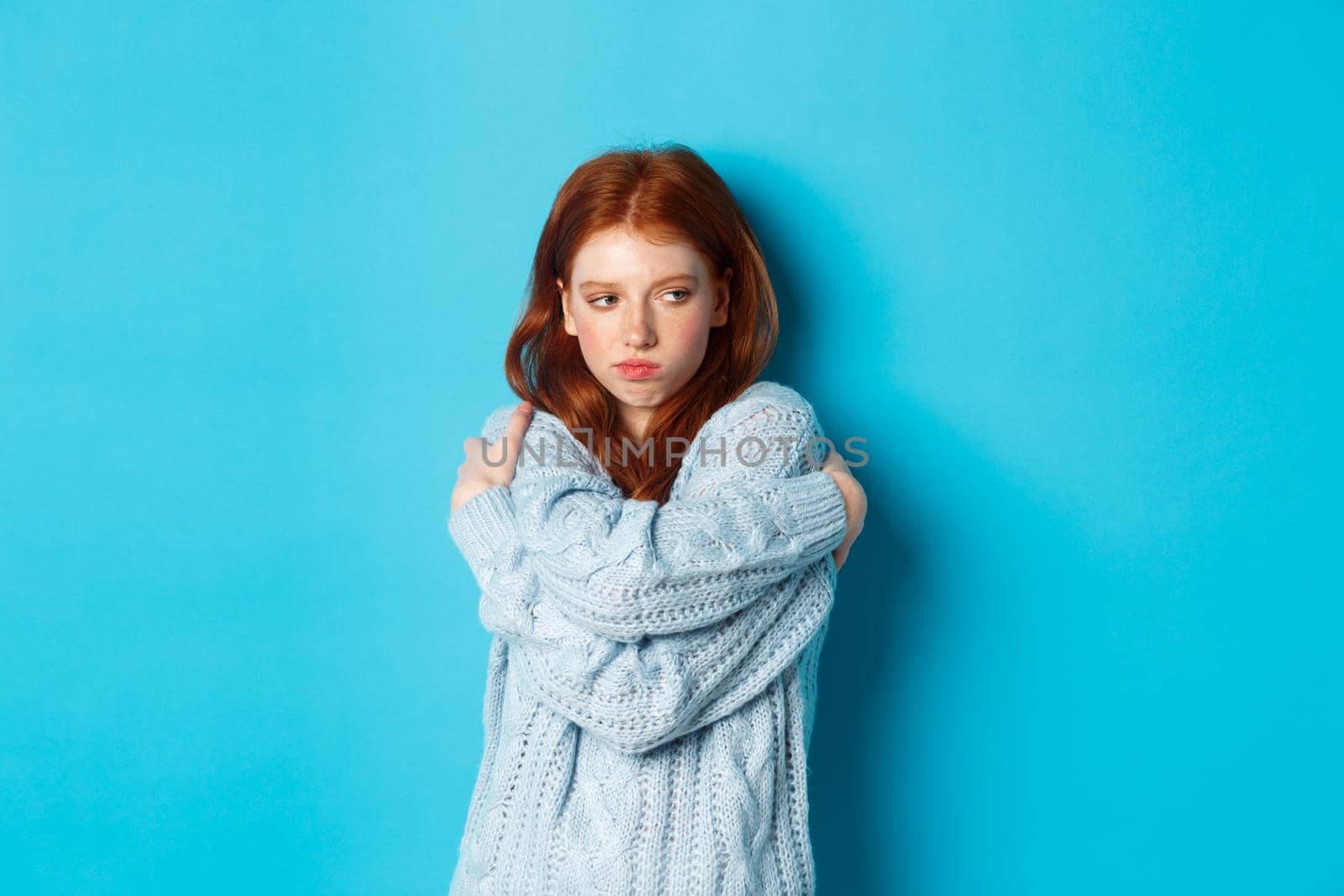 Silly and cute redhead girl pucker lips and looking offended, hugging herself and looking away offended, sulking and being upset, standing over blue background.