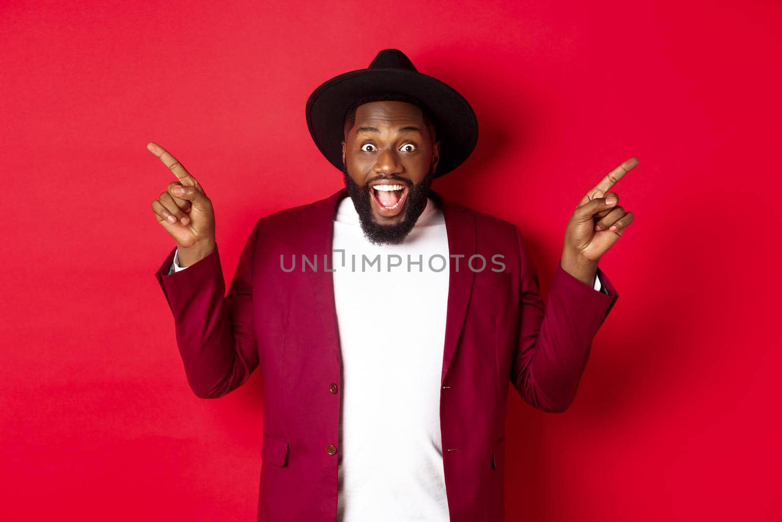 Winter holidays and shopping concept. Cheerful Black man smiling and showing two promo, pointing fingers sideways at copy spaces, red background.