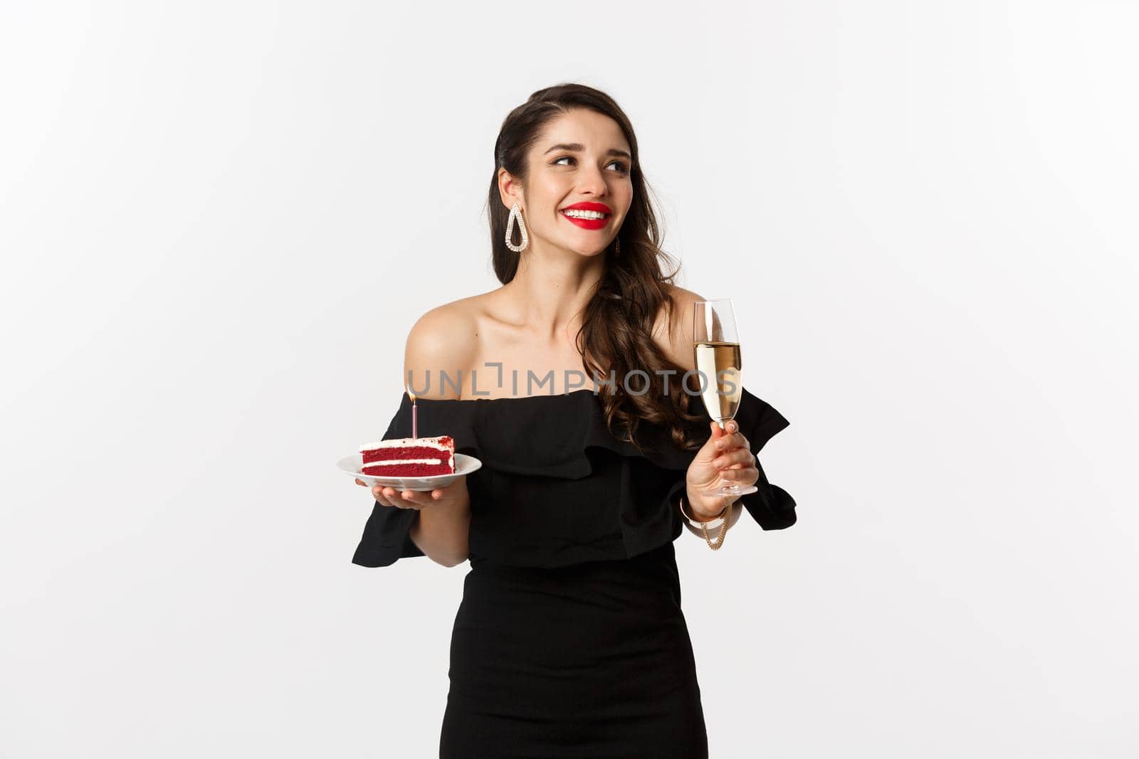 Celebration and party concept. Fashionable woman holding birthday cake with candle and drinking champagne, smiling and looking aside, standing over white background.