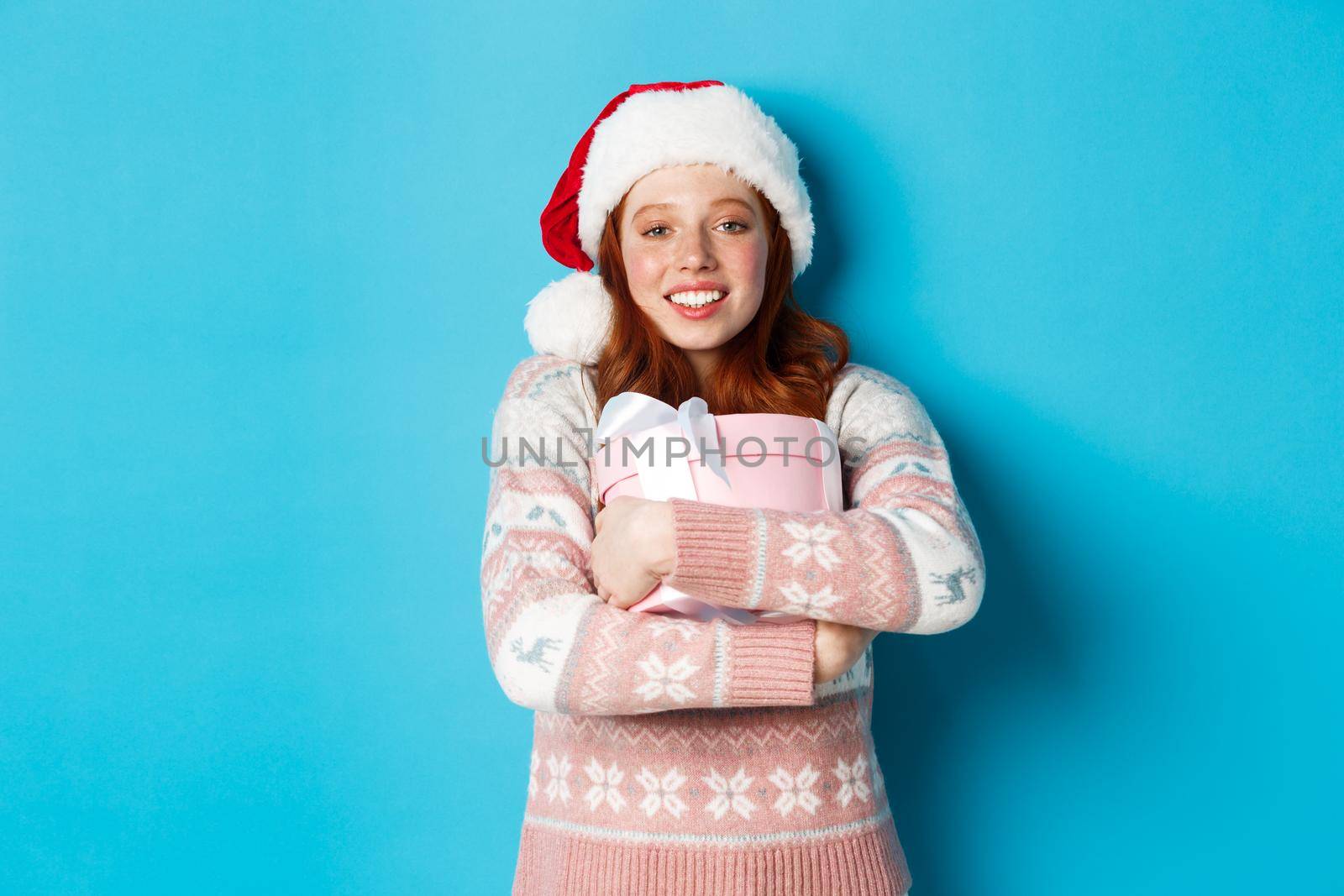 Winter and celebration concept. Dreamy redhead girl in santa hat hugging her christmas gift, smiling happy, standing over blue background.