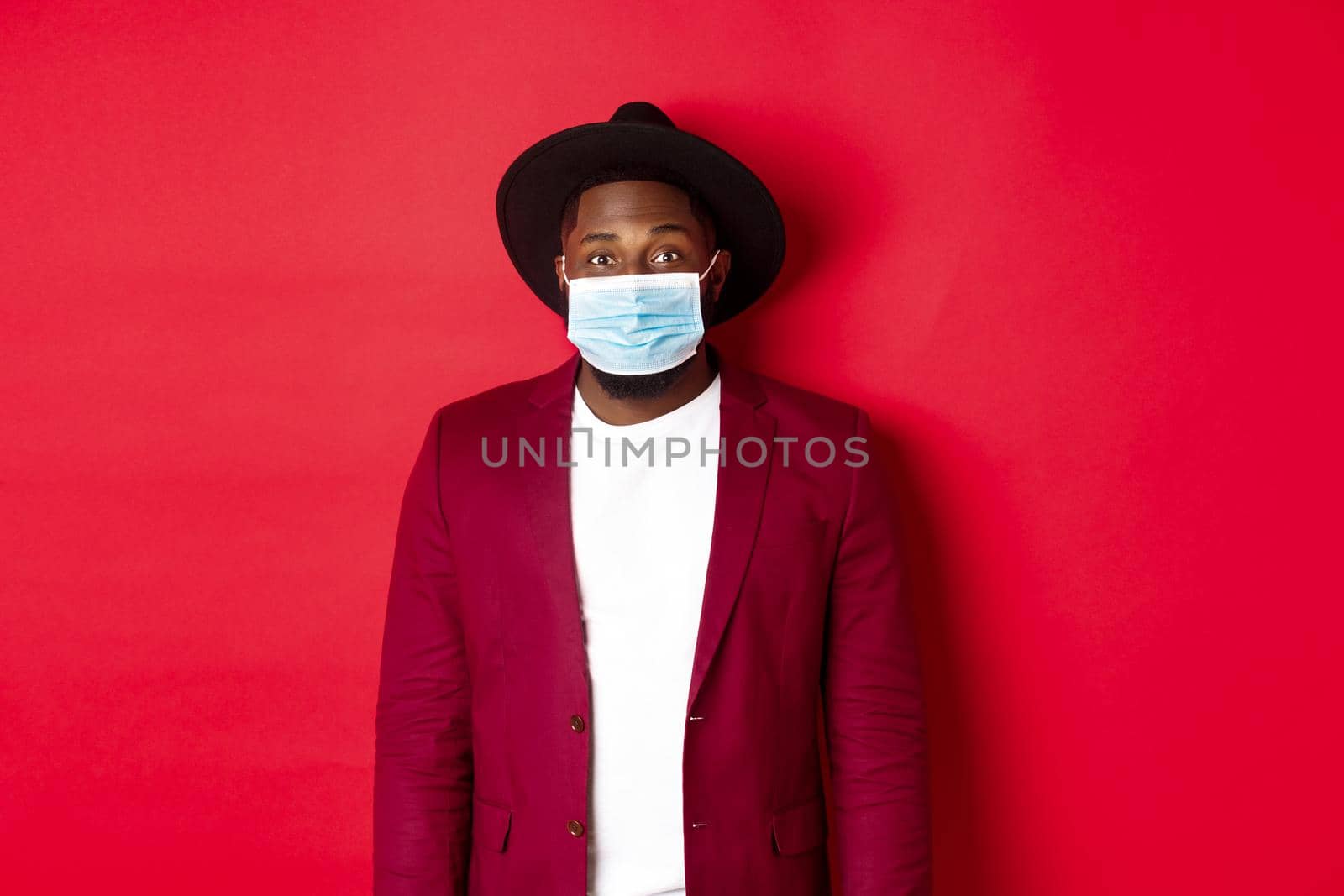 Covid-19 and fashion concept. Handsome african american man in stylish blazer and hat, wearing medical mask from coronavirus, standing over red background.