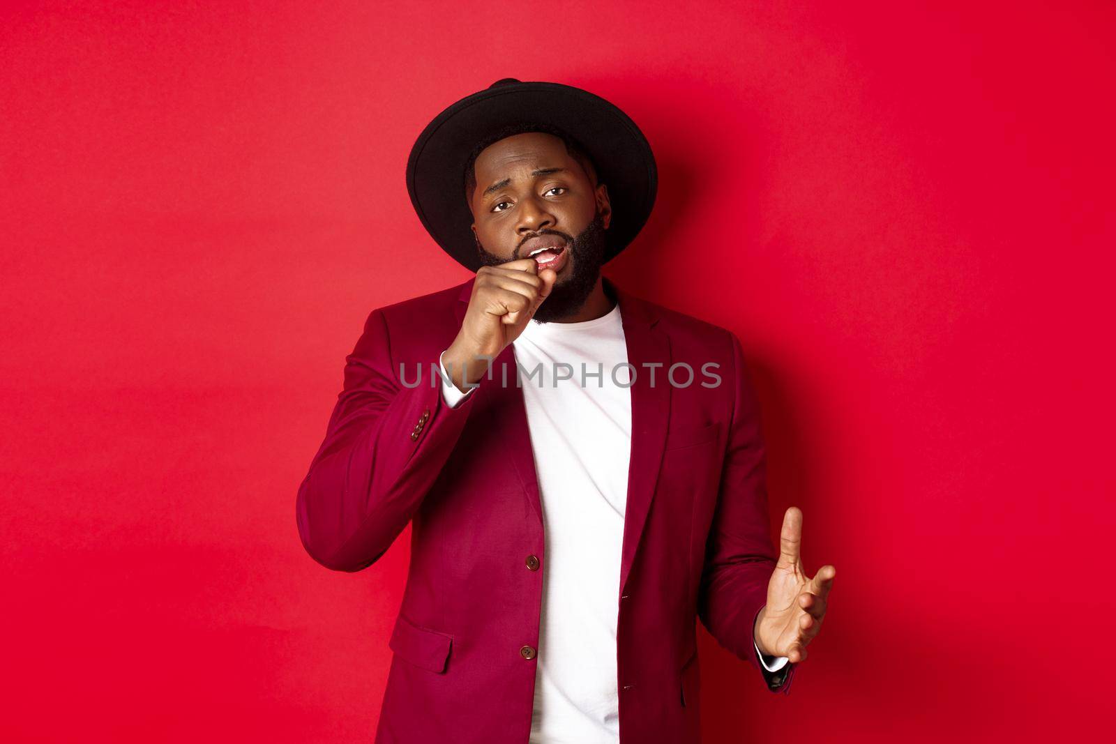 Christmas shopping and people concept. Handsome Black male model in party outfit singing in invisible microphone, looking passionate at camera, red background.