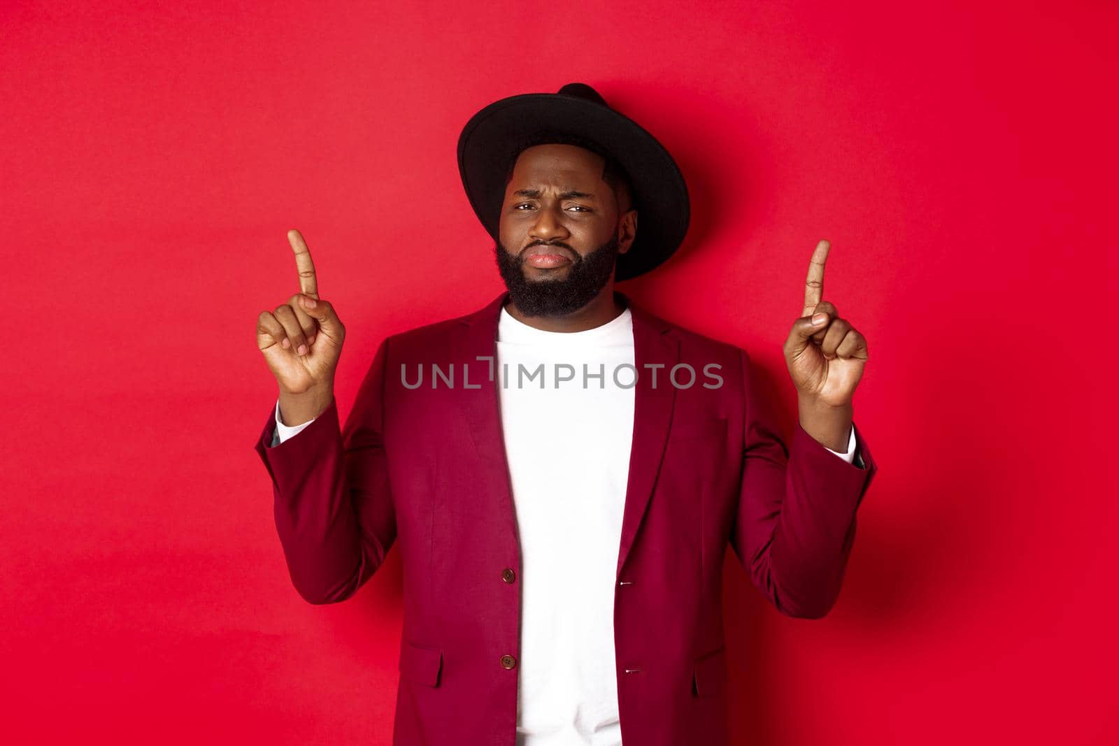 Winter holidays and shopping concept. Skeptical Black man grimasing and showing dislike, pointing fingers up at bad offer, standing over red background.