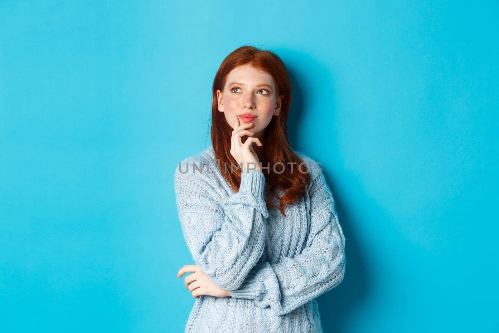 Thoughtful cute woman with red hair, looking upper left corner logo and thinking, imaging something, standing over blue background.