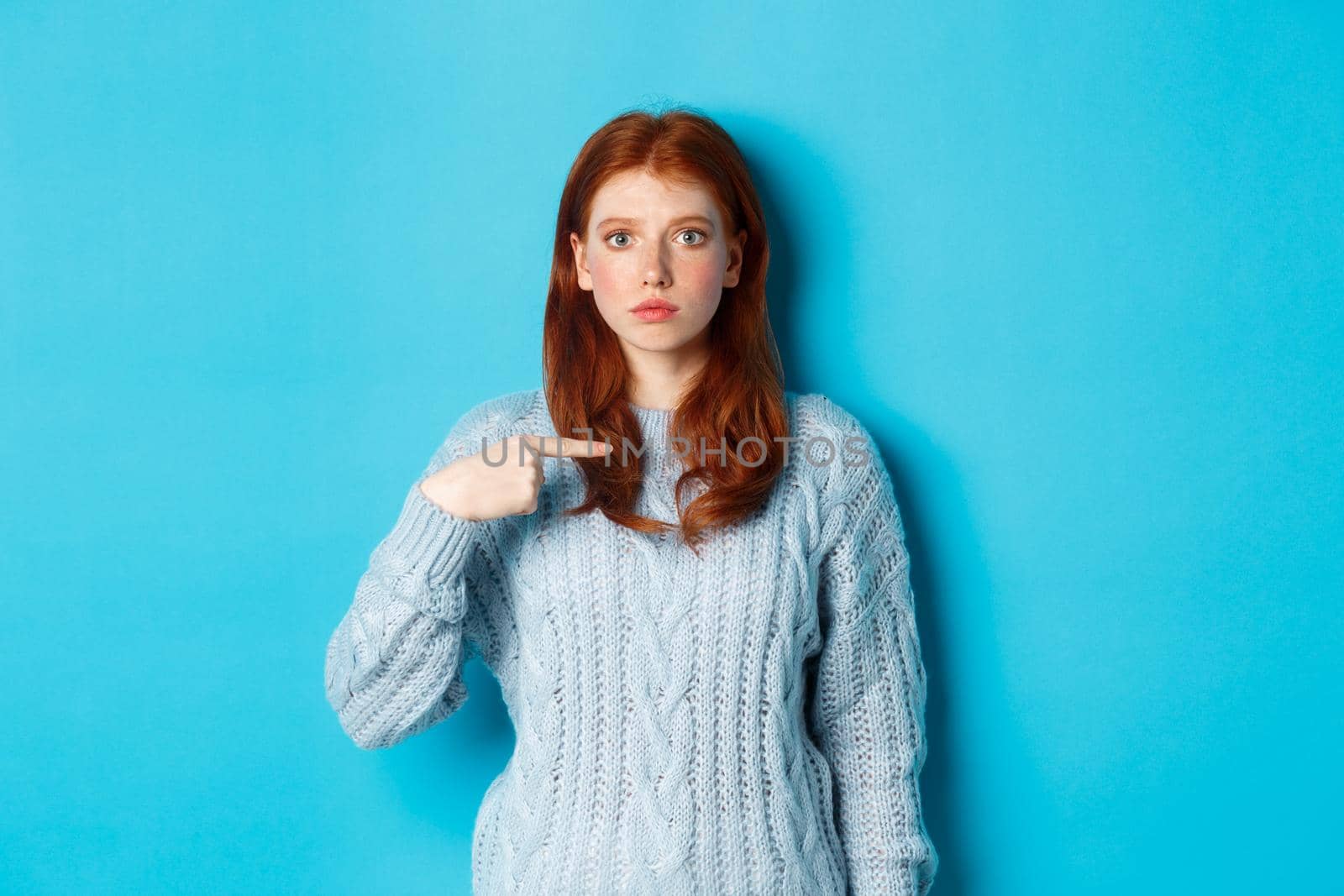 Nervous and confused redhead girl pointing at herself, standing in sweater against blue background.